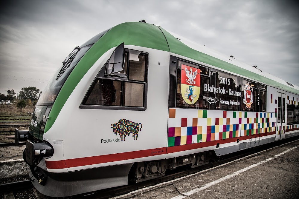 Pociąg stoi na torach. Pojazd ma zielony przód, jego bok jest w kolorowe kwadraty, z których składa się żubr - symbol województwa podlaskiego, który widoczny jest na boku pociągu