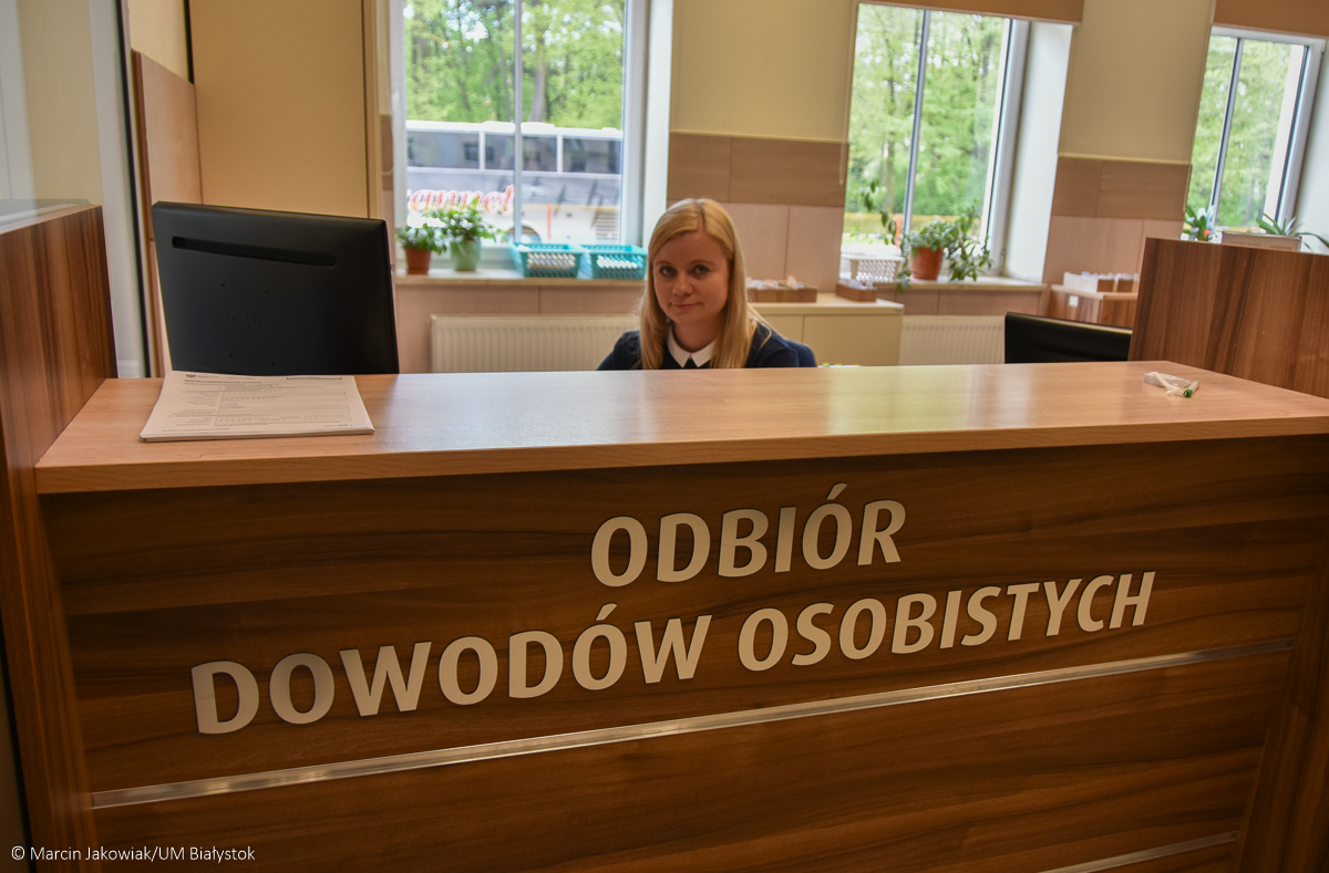 Kobieta o blond włosach siedzi za drewinanym blatem, na którego przedniej ściance jest napisane: Odbiór dowodów osobistych