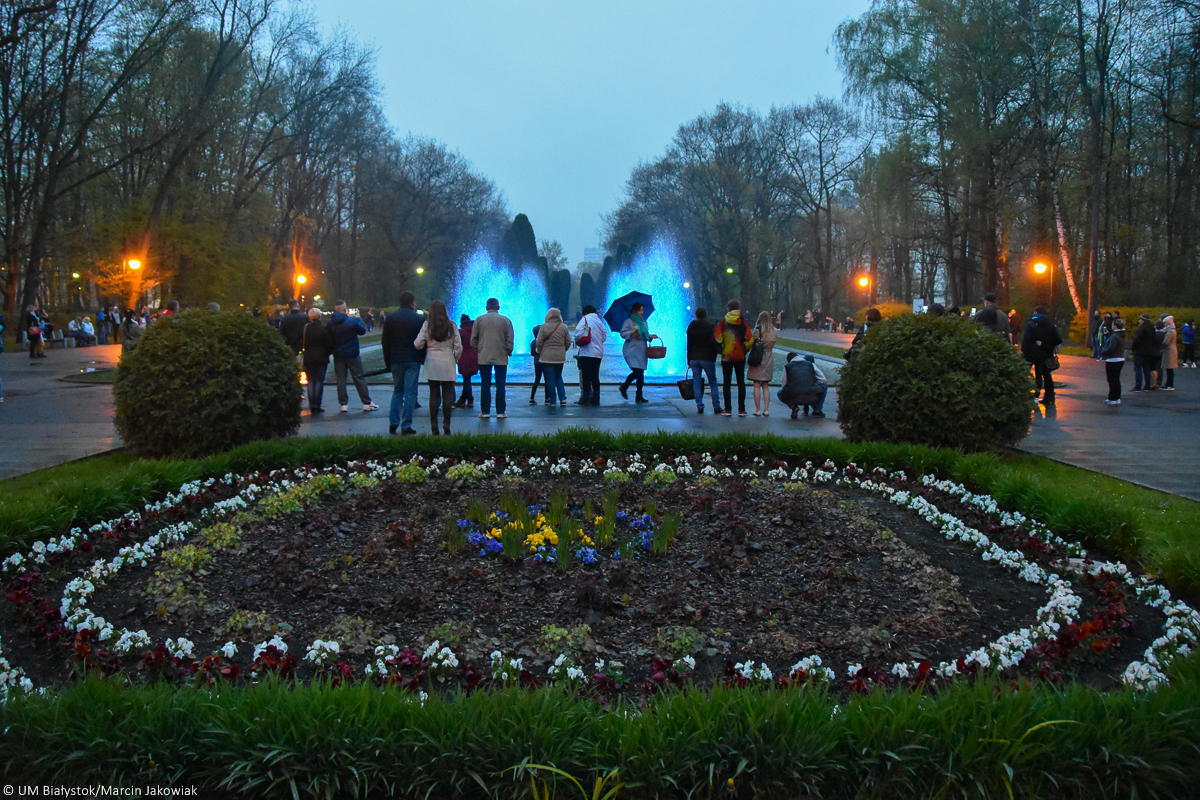Park Plany w Białymstoku. Na pierwszym planie jest kompozycja z posadzonych kwiatów. W oddali widoczne są kolorowe fontanny, przy których stoją ludzie