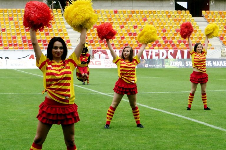 Czirliderki żółto-czerwonych stoją na płycie boiska. W rękach trzymają pompony w kolorach klubu