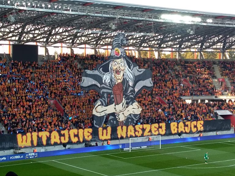 Widać fragment zielonej murawy oraz trybun, gdzie umieszczono oprawę stadionową. Przedstawia ona wiedźmę, pod którą jest napis: Witajcie w naszej bajce