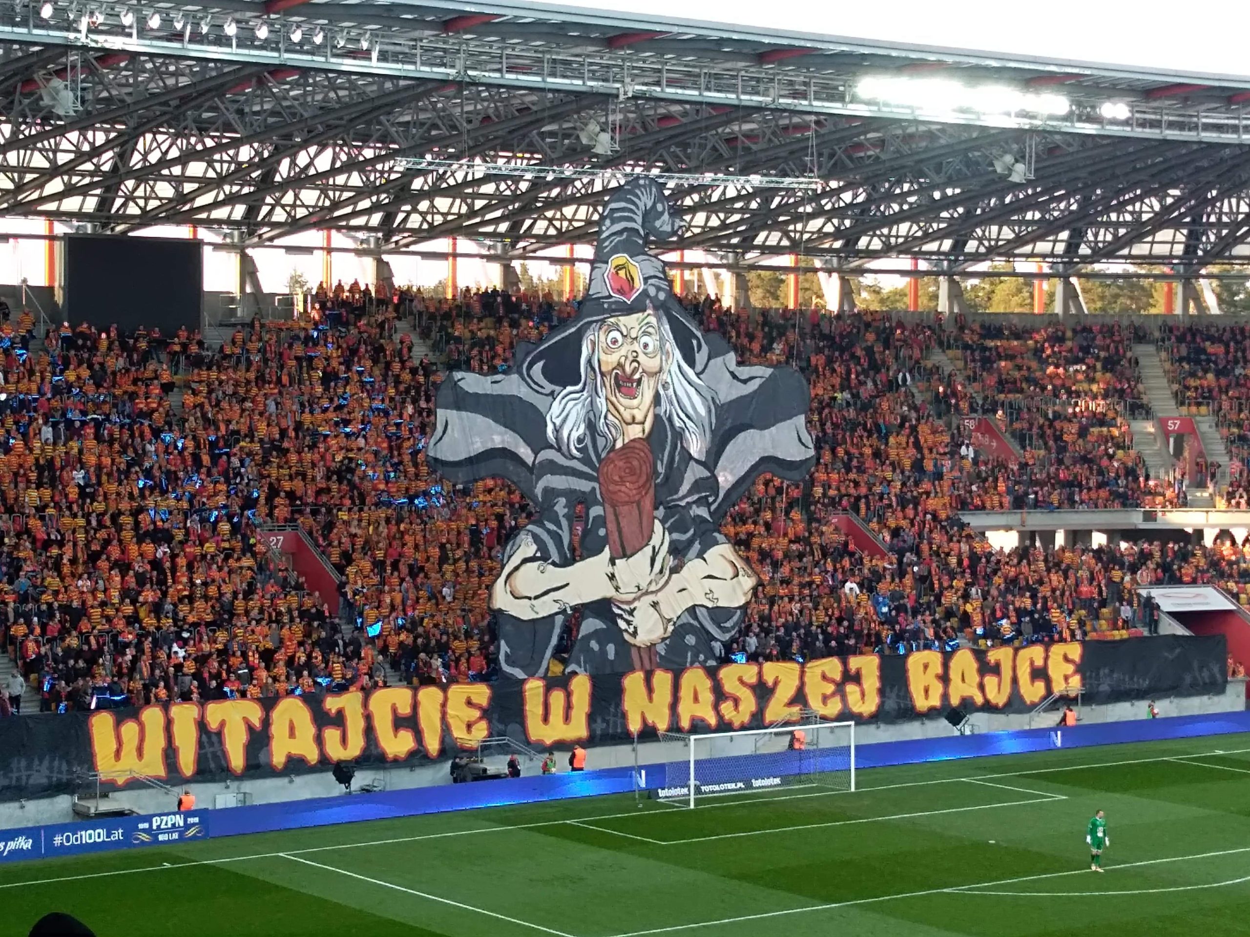 Widać fragment zielonej murawy oraz trybun, gdzie umieszczono oprawę stadionową. Przedstawia ona wiedźmę, pod którą jest napis: Witajcie w naszej bajce