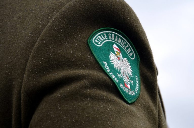 Zielona naszywka z godłem Polski i napisem Straż Graniczna przyszyta do brązowego płaszcza funkcjonariusza. Widoczny jest tylko fragment przedramienia