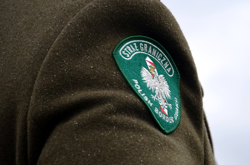 Zielona naszywka z godłem Polski i napisem Straż Graniczna przyszyta do brązowego płaszcza funkcjonariusza. Widoczny jest tylko fragment przedramienia