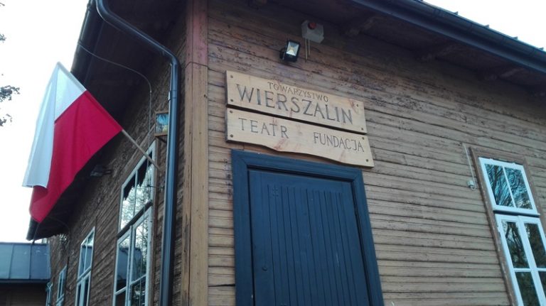 Fragment drewnianego budynku. Uchwycony jest narożnik, z lewej ścianie jest okno, nad którym wisi flaga biało-czerwona flaga Polski. Na prawej ścianie są ciemnobrązowe drzwi, nad którymi wisi drewniana tabliczka z napisem: Towarzystwo Wierszalin, Teatr Fundacji, dalej na ścianie jest też okno w jasnozielonej oprawie