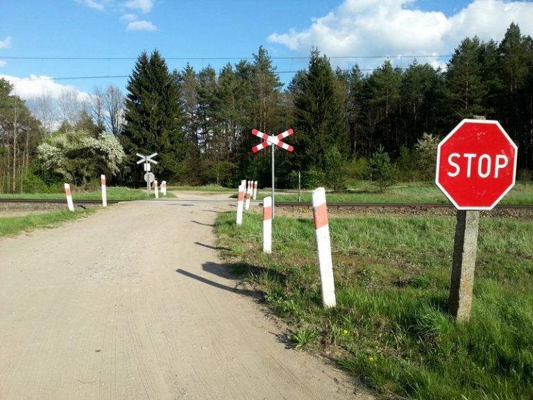 Droga prowadząca do przejazdy kolejowego. Po prawej stronie stoi czerwony znak z białym napisem: STOP w oddali widoczny jest krzyż św. Andrzeja