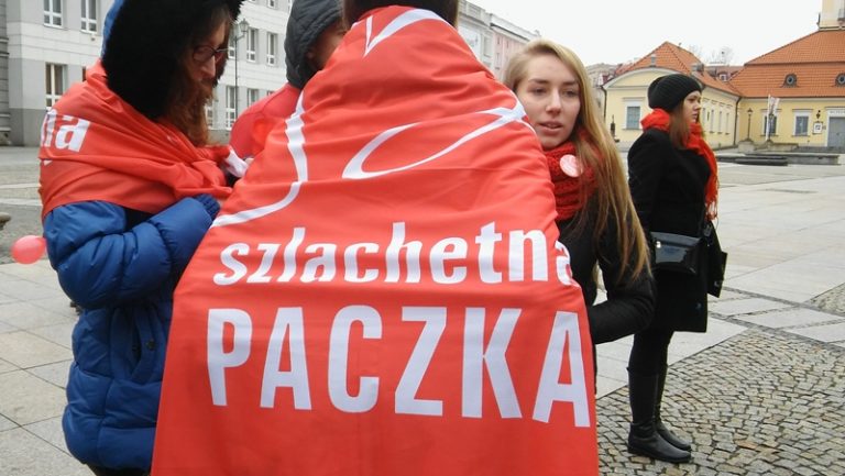 Wolontariusze stoją na Rynku Kościuszki w Białymstoku. Osoba na pierwszym planie jest odwrócona plecami, jest owinięta w flagę Szlachetnej Paczki