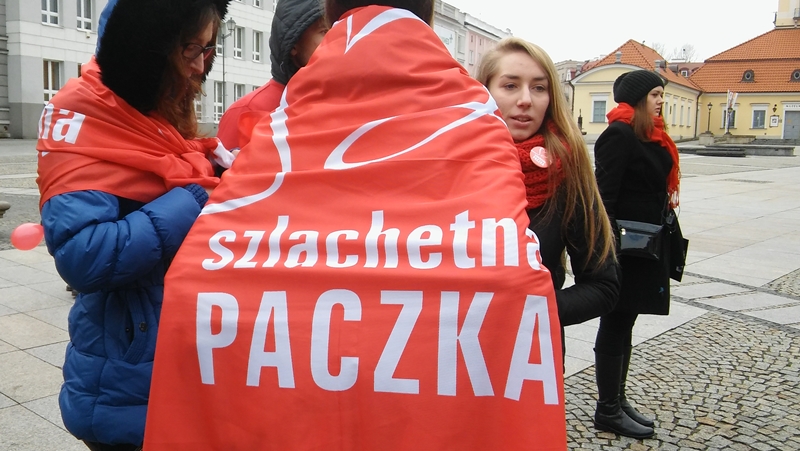 Wolontariusze stoją na Rynku Kościuszki w Białymstoku. Osoba na pierwszym planie jest odwrócona plecami, jest owinięta w flagę Szlachetnej Paczki