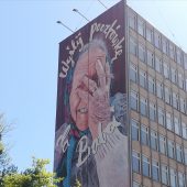 Wysoki budynek, na którego bocznej ścianie znajduje się mural. Malowidło przedstawia straszą panię w chustce na głowie, która dłonią zasłania twarz. Na muralu znajduje się napis: Wyślij pocztówkę do babci.