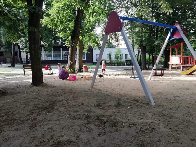 Na piachu stoi huśtawka, w oddali przy drzewie siedzą ludzie