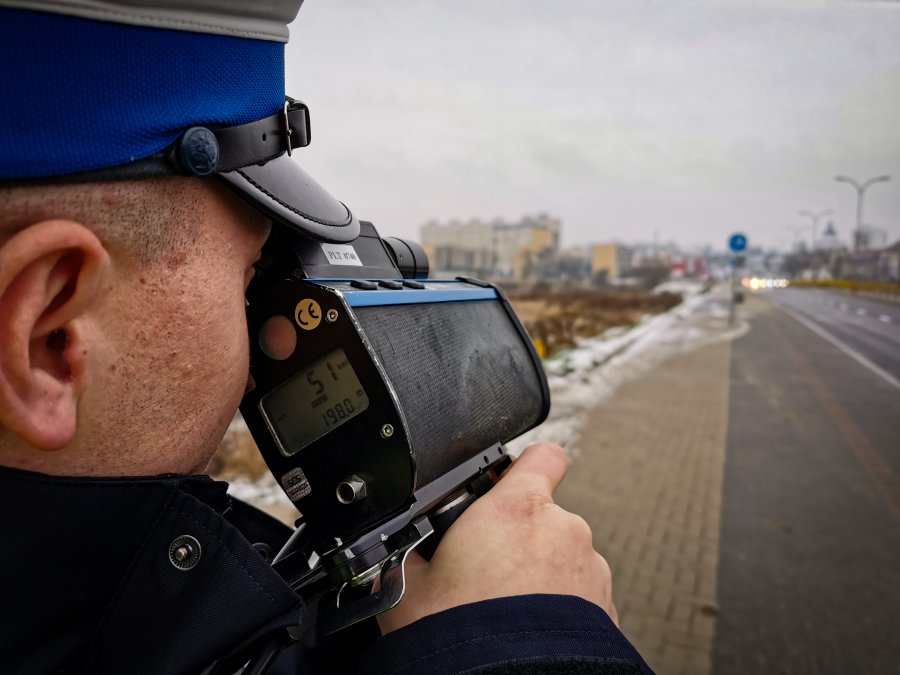 Policjant trzyma przy twarzy prędkościomierz, po prawej stronie widać ulicę