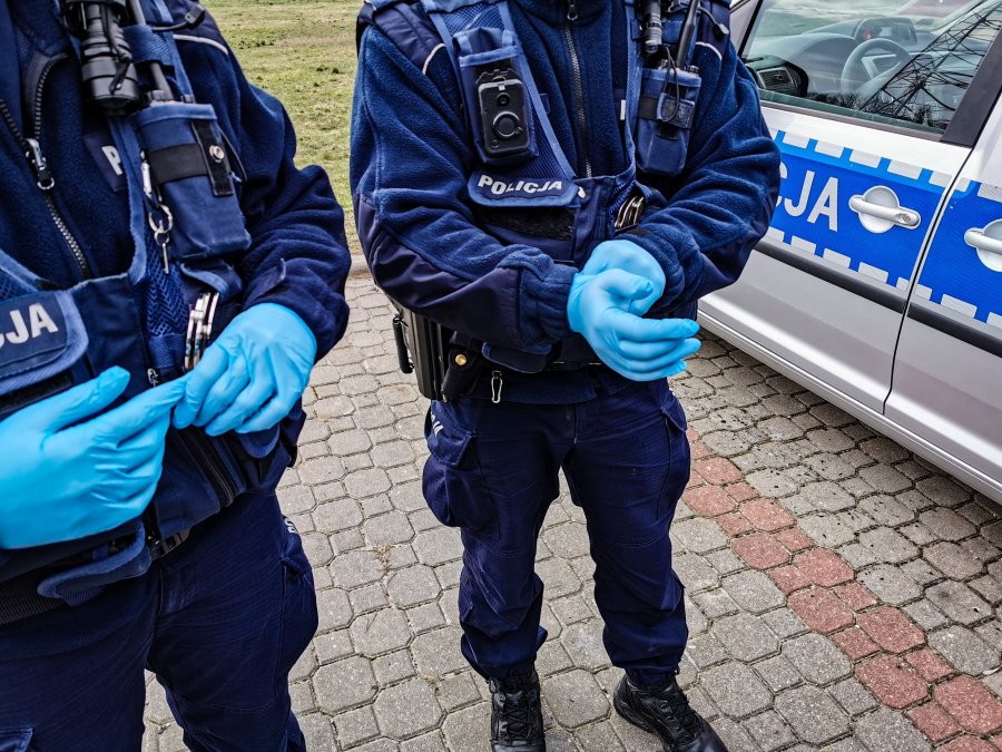 Dwaj policjanci stoją przy radiowozie, na rękach mają jednorazowe rękawiczki