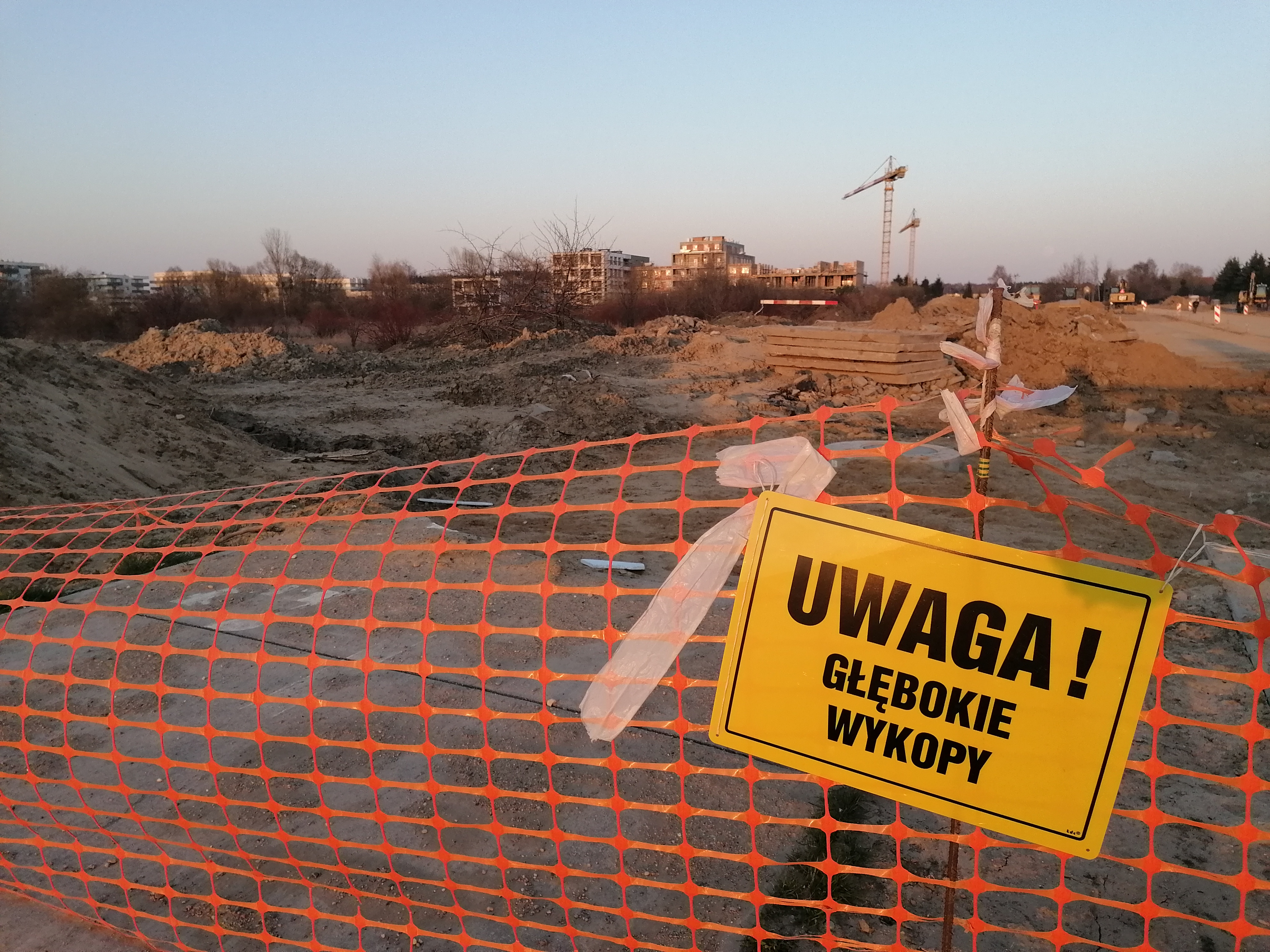 na ogrodzeniu z siatki wisi znak Uwaga głębokie wykopy, za siatką miejsce pod budowę