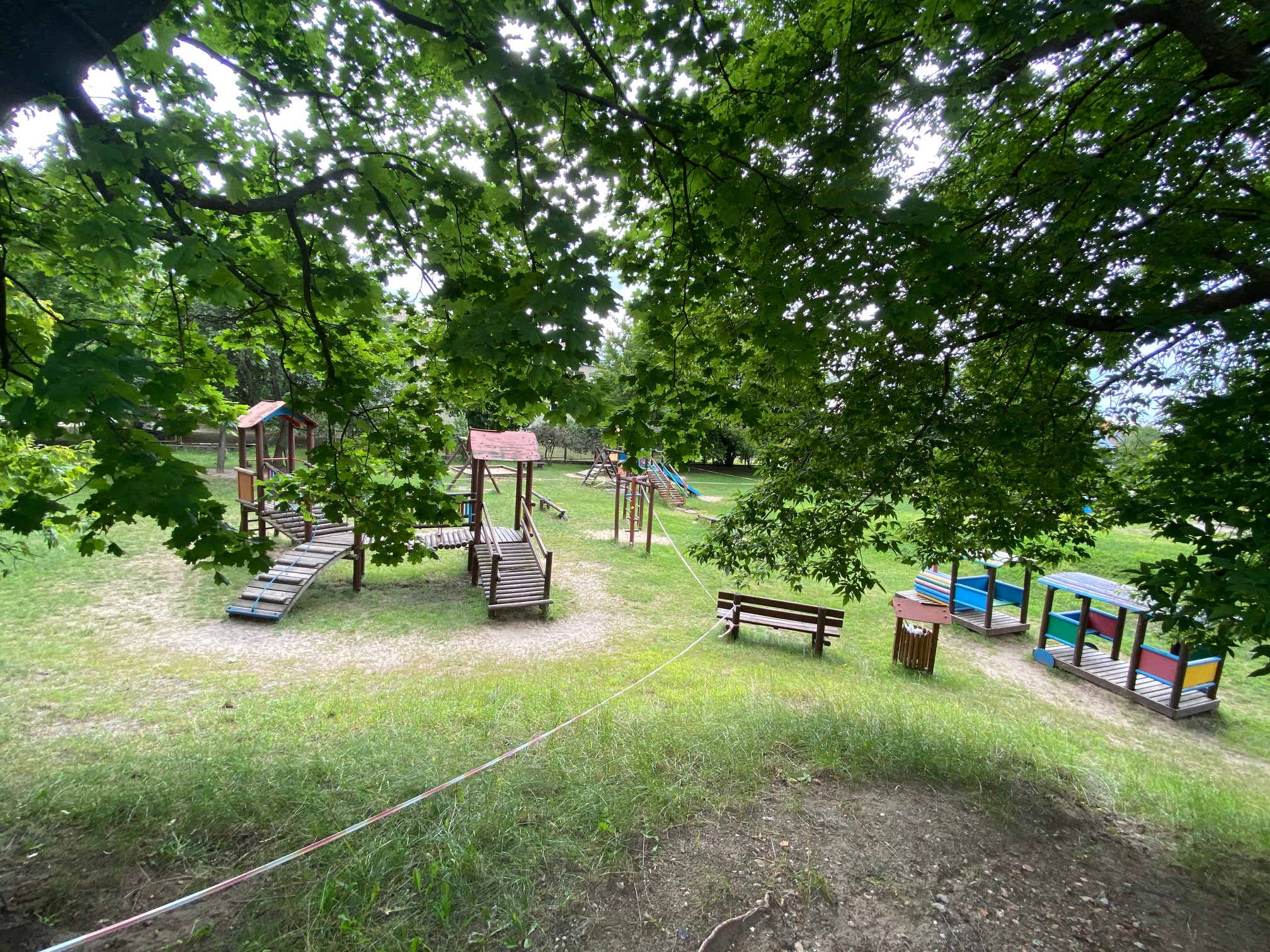 Plac zabaw położony wśród zieleni. W oddali widać drabinki, ławki