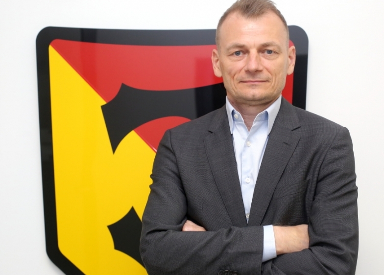 Na zdjęciu trener Jagiellonii Białystok, Bogdan Zając. Mężczyzna stoi z założonymi rękami, za jegeo plecami jest duże logo białostockiego zespołu.