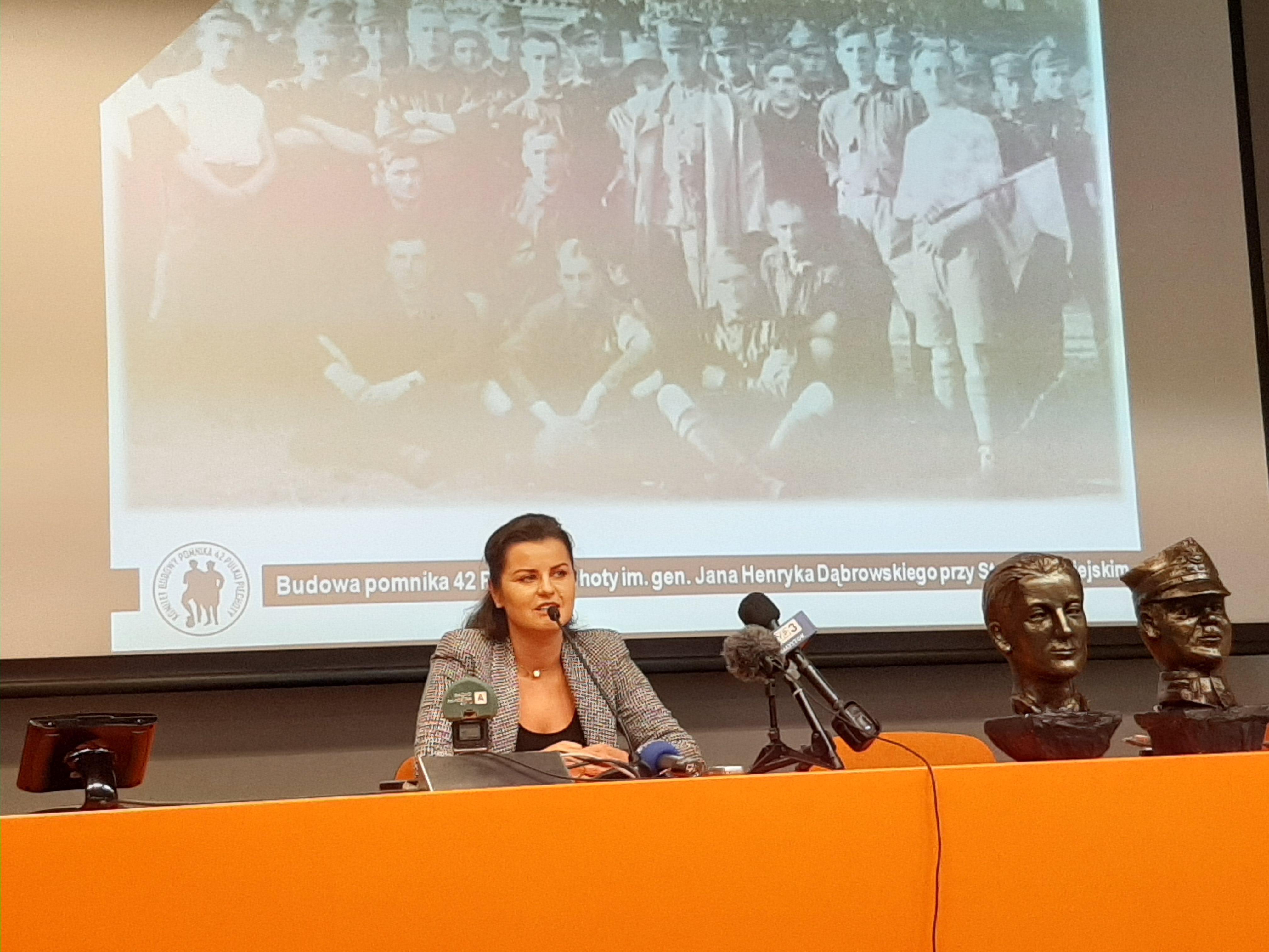 Sala konferencyjna Stadionu Miejskiego w Białymstoku. Przy stole siedzi wiceprezes Jagiellonii Agnieszka Syczewska, która mówi o budowie pomnika. Za nią widoczne jest zdjęcie 42. Pułku Piechoty
