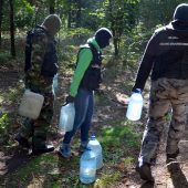 Funkcjonariusze KAS stoją na terenie leśnej bimbrowni. Na miejscu jest kadź zawierająca 300 litrów fermentującego zacieru, chłodnica i kocioł, a także doły z wodą wykorzystywaną w wytwarzaniu bimbru oraz rozlewisko