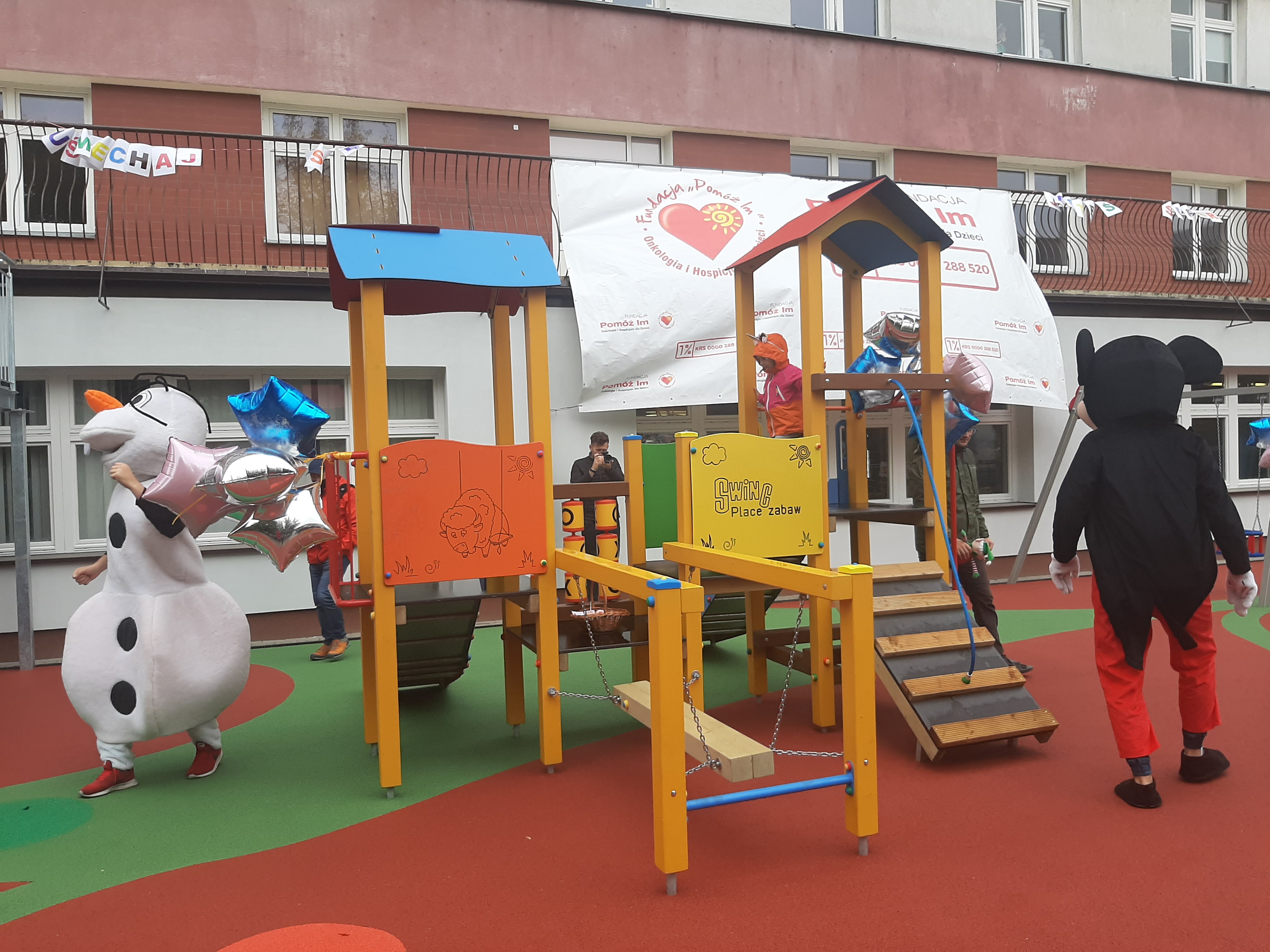 Plac zabaw przed oddziałem onkologii UDSK. Dzieci biegają po drabinkach. Z lewej strony jest chodząca maskotka bałwan, z prawej zaś tyłem odwrócona Myszka Miki