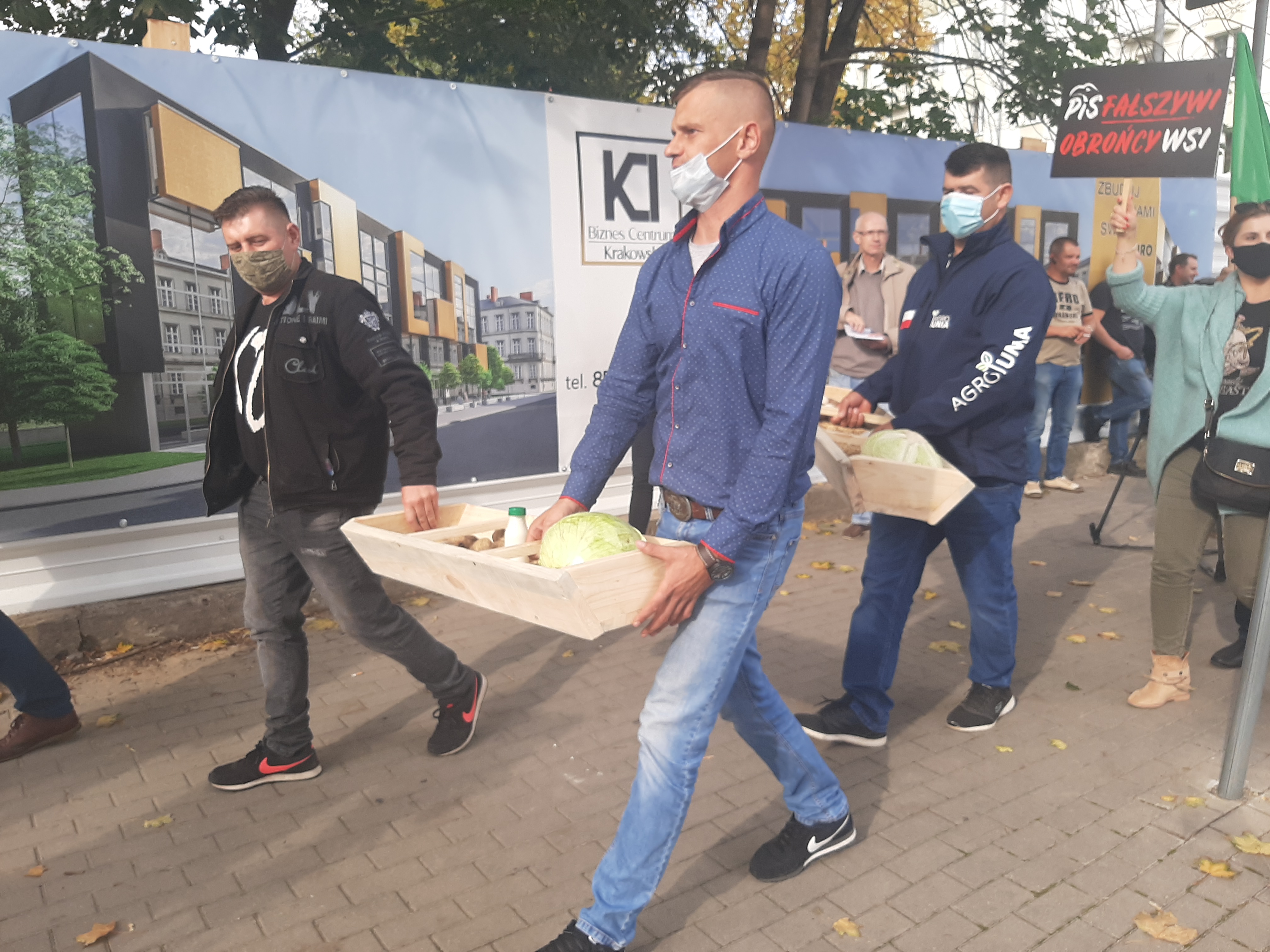 Na pierwszym planie znajduje się dwóch mężczyzn, którzy na drewnianej tacy niosą kapustę, ziarna, mleko, jajka. Za nimi idą kolejne osoby z żywnością. Obok nich jest kobieta z transparentem, na którym jest napisane PiS Fałszywi Obrońcy Wsi