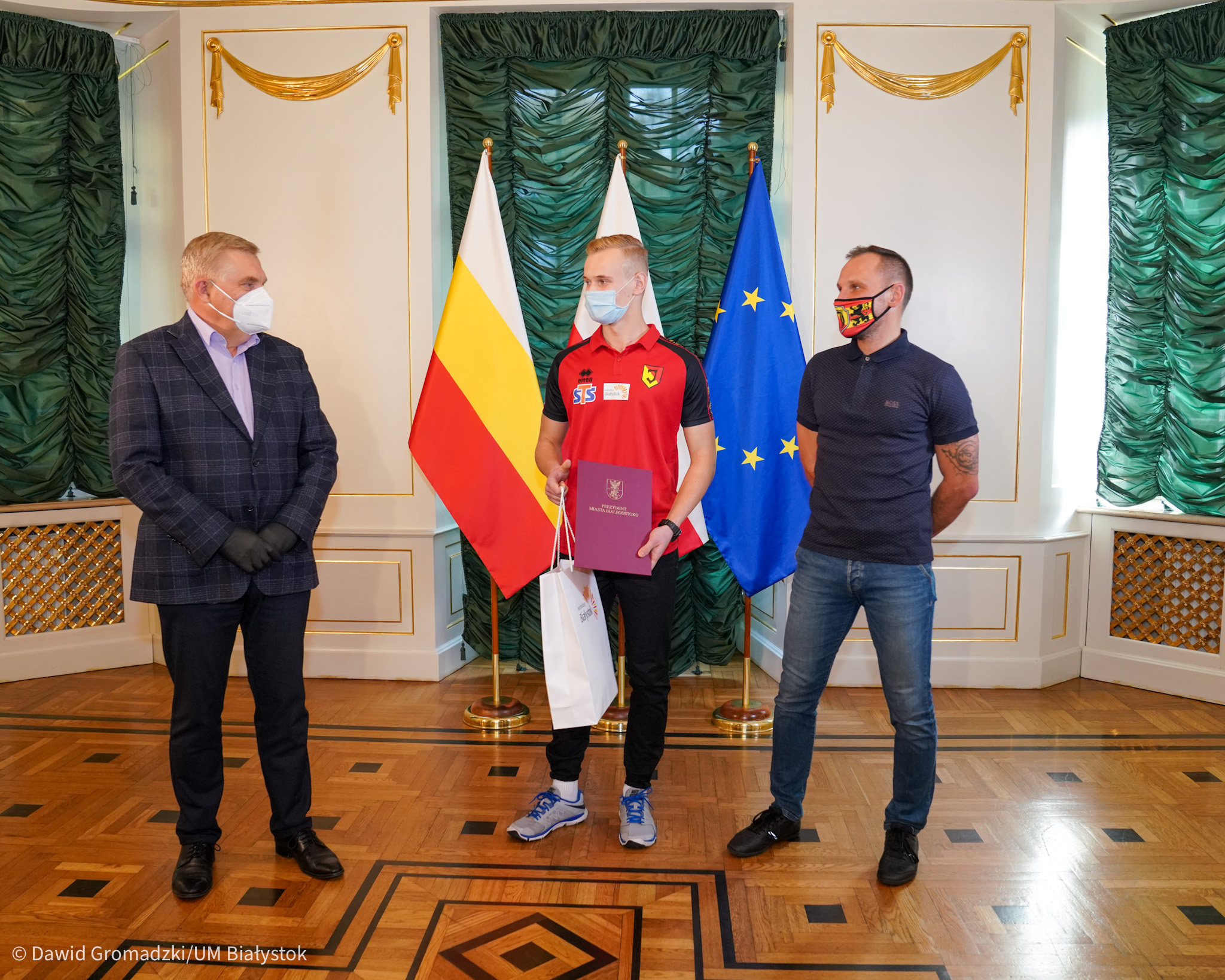 W sali Pałacyku Gościnnego w Białymstoku, po lewej stronie stoi prezydent miasta ubrany w ciemny garnitur, po środku nastolatek ubrany w koszulkę Jagiellonii i ciemne spodnie, a po lewej stronie znajduje się mężczyzna ubrany w czarną koszulkę oraz jeansy. Mężczyźni patrzą na siebie