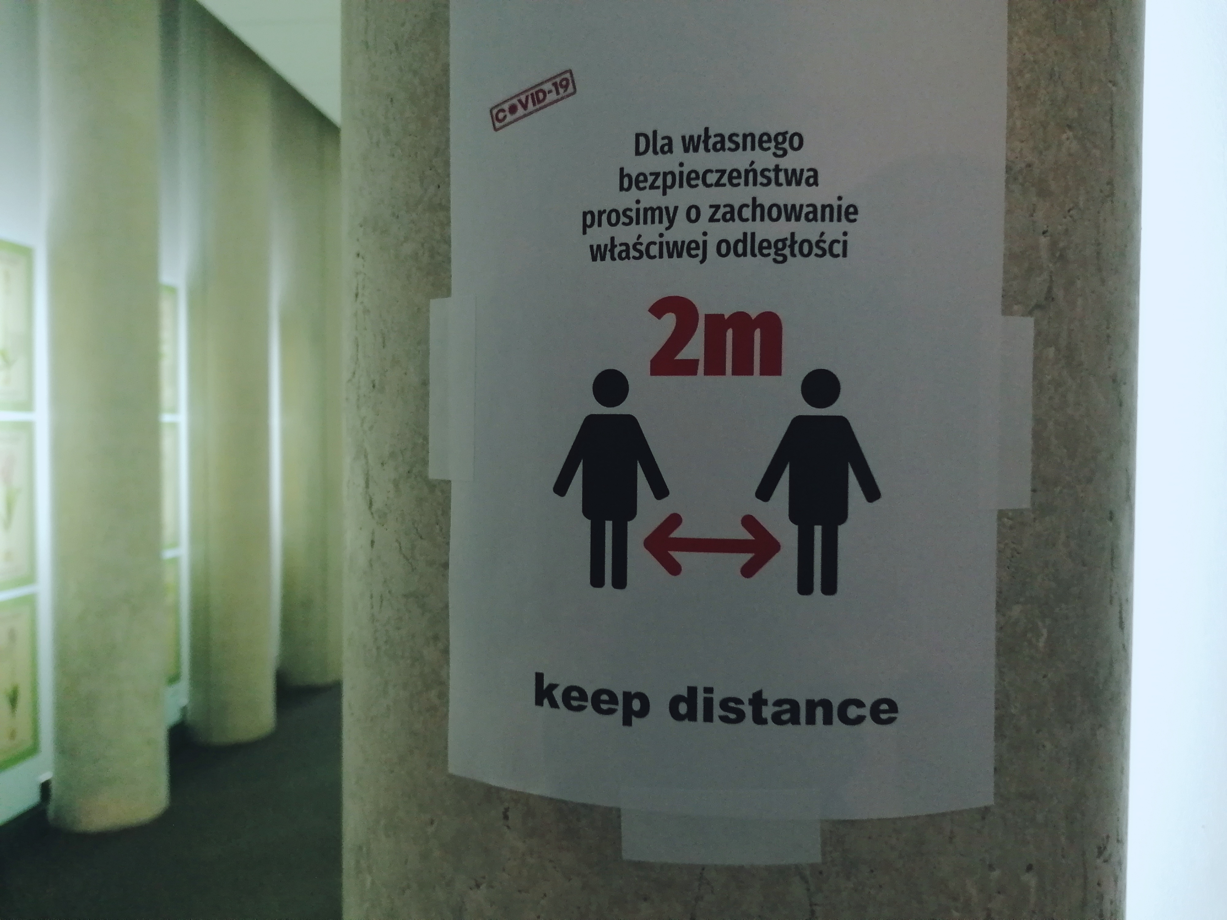 Na słupie w korytarzu wisi plakat przypominający o obowiązku zachowania dystansu społecznego.