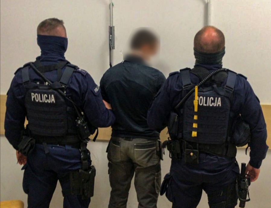 2 policjantów w mundurach, którzy trzymają zatrzymanego mężczyznę.