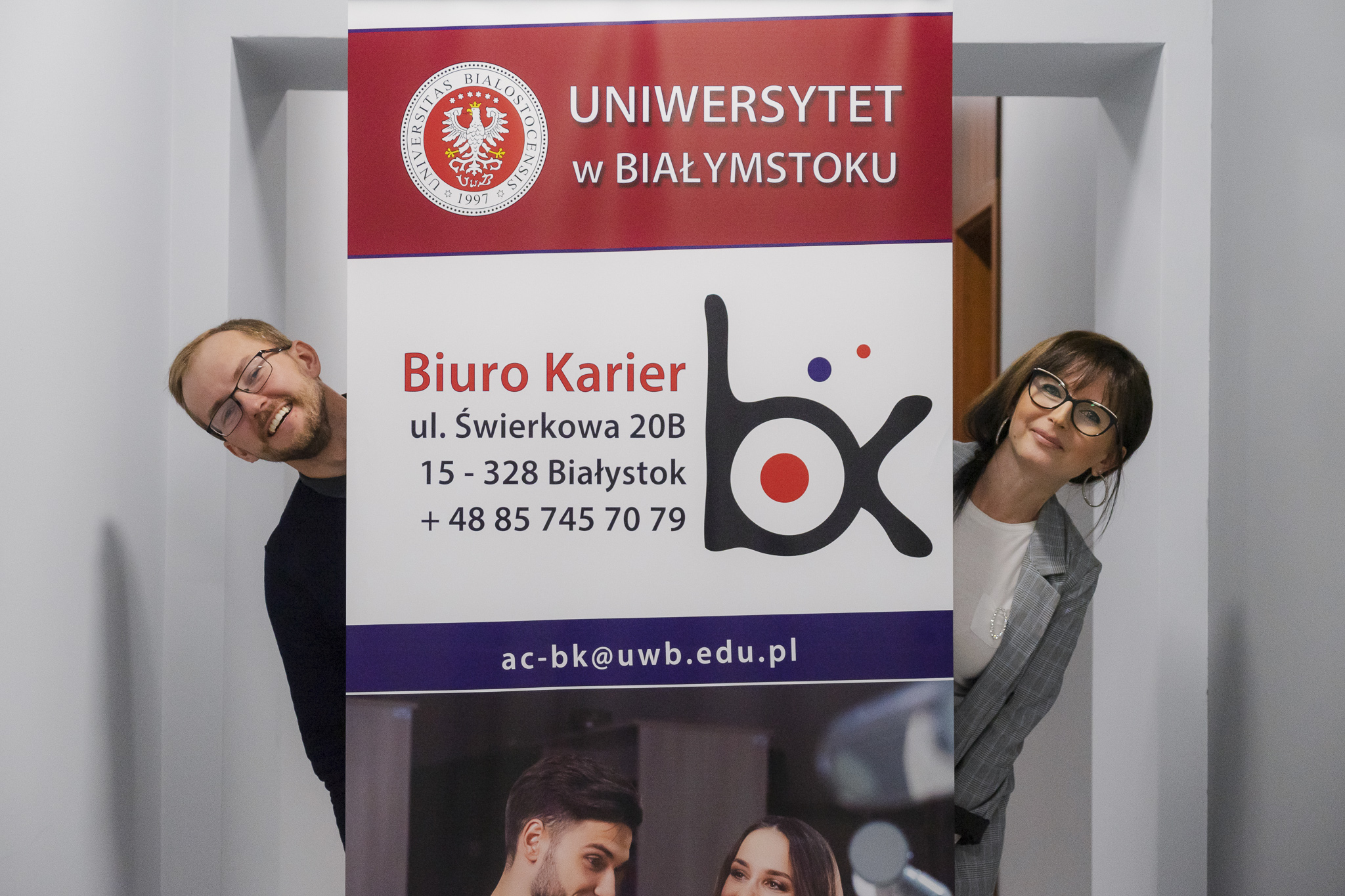 Mężczyzna i kobieta wyglądają zza roll-upu reklamującego Biuro Karier Uniwersytetu w Białymstoku.