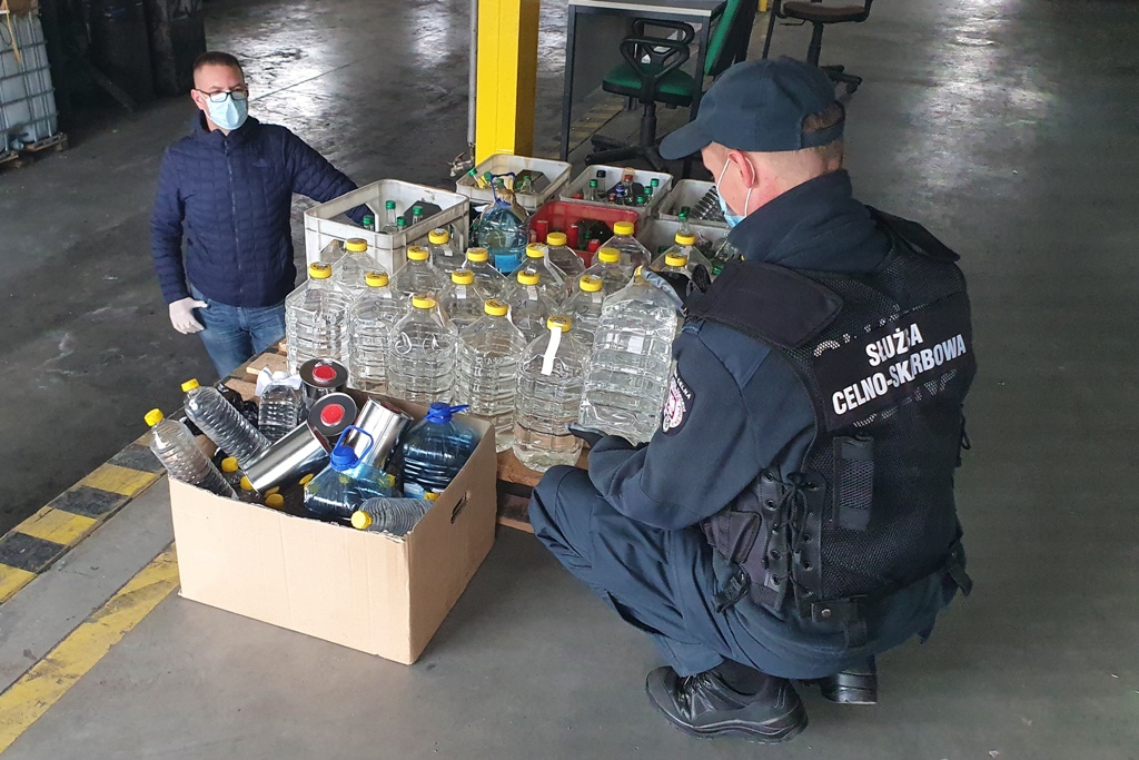 Funkcjonariusz Krajowej Administracji Skarbowej kuca przy plastikowych butelkach wypełnionych alkoholem. Darowiznę od funkcjonariusza odbiera mężczyzna . Przekazanie odbywa się w hali