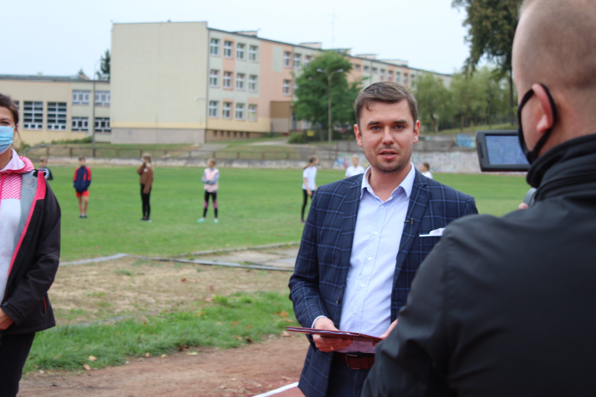 Na terenie boiska szkolnego stoi burmistrz Wasilkowa. W oddali dzieci grają w piłkę.