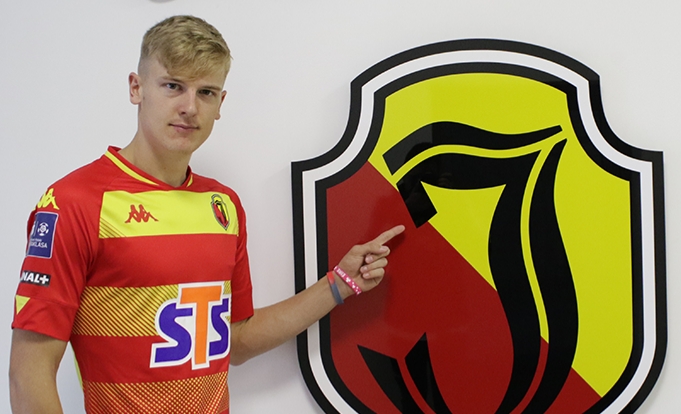 Piłkarz Jagiellonii Białystok, Kris Twardek stoi w klubowej, żółto-czerwonej koszulce i wskazuje palcem na duże logo drużyny znajdujące się na ścianie.