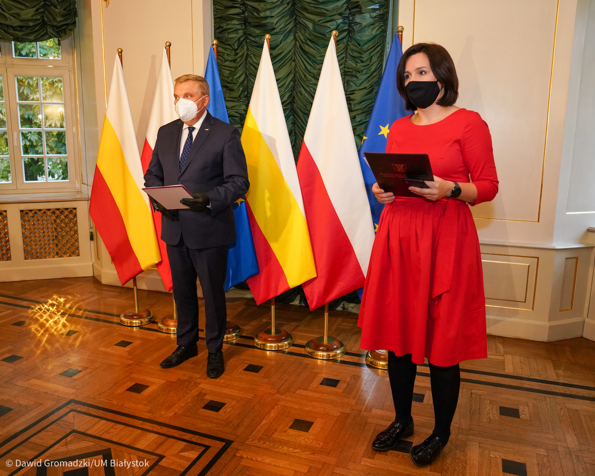 Po lewej stronie stoi prezydent Białegostoku . Za jego plecami są ustawione flagi Polski oraz Białegostoku. Po prawej stronie w czerwonej sukience stoi rzeczniczka prezydenta. Sytuacja ma miejsce w sali Pałacyku Gościnnego