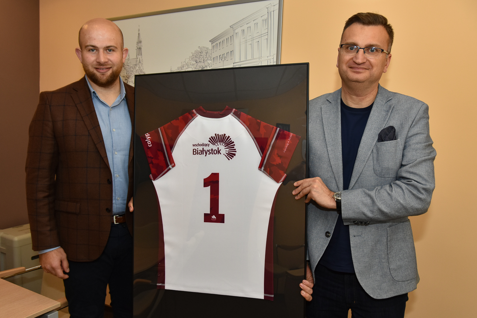 Po lewej stronie stoi mężczyzna ubrany w granatowe spodnie, jasno niebieską koszulę i bordową marynarkę. Po prawej stronie stoi zastępca prezydenta Białegostoku ubrany w ciemną koszulkę i szarą marynarkę. Panowie trzymają białą z czerwonymi rękawami koszulkę do gry w rugby. T-shirt to prezent od drużyny rugby dla wiceprezydenta