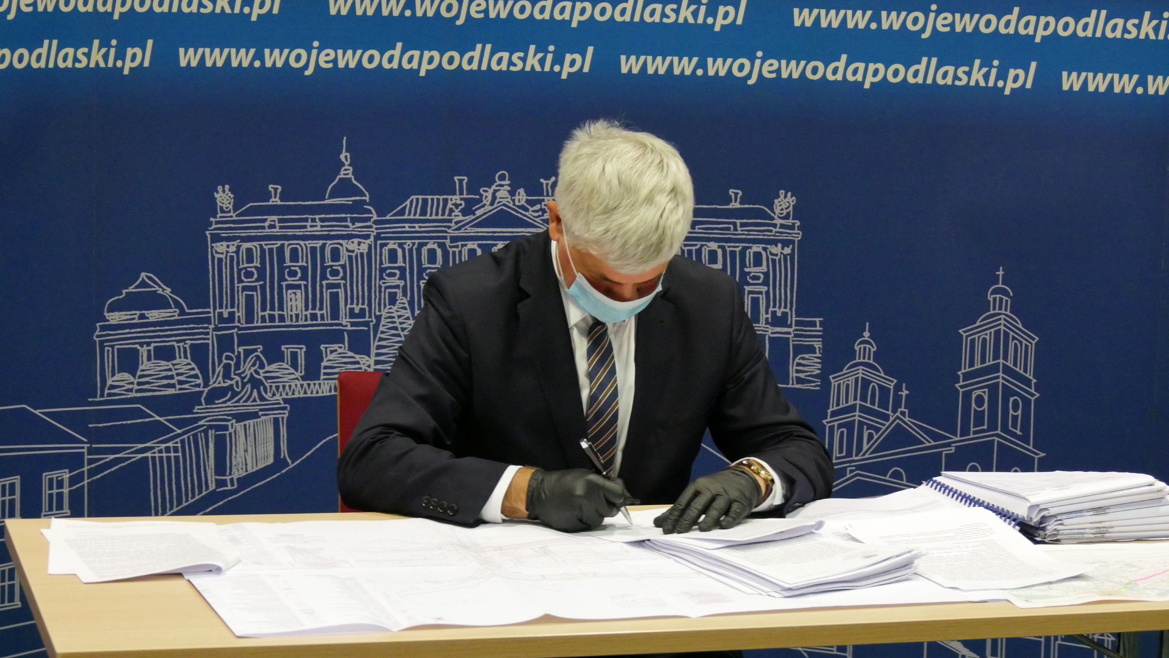 Wojewoda pochylony nad planami rozbudowy tras kolejowych podpisuje pozwolenie na kolejną rozbudowę odcinka. Wojewoda jest ubrany w czarny garnitur, za nim niebieska ścianka promocyjna Podlaskiego Urzędu Wojewódzkiego w Białymstoku