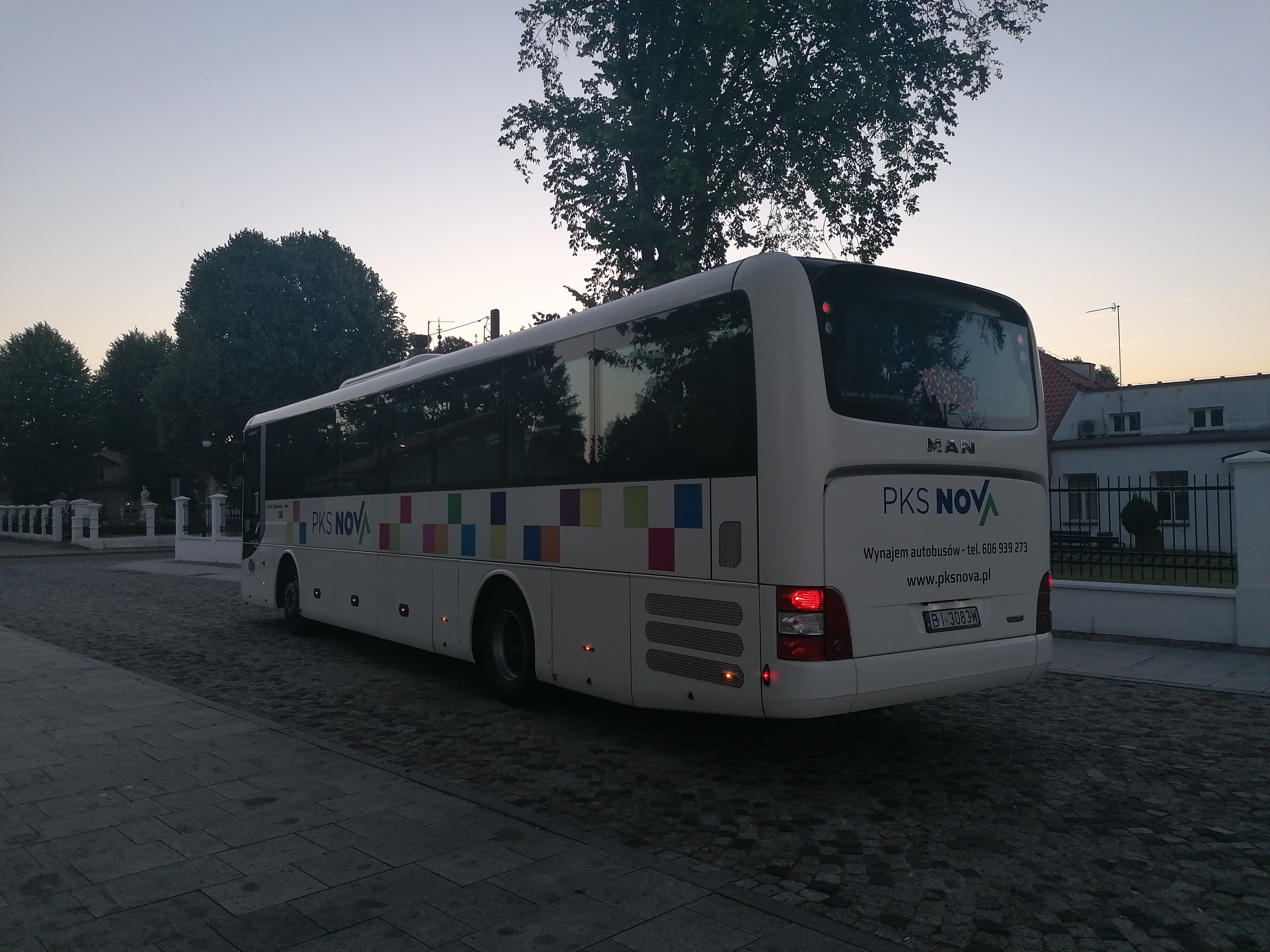 Na brukowanej ulicy stoi biały, w kolorowe kwadraty autobus, typu autokar. Pojazd stoi tyłem, widoczna jest tylna szyba i jego boczna ściana
