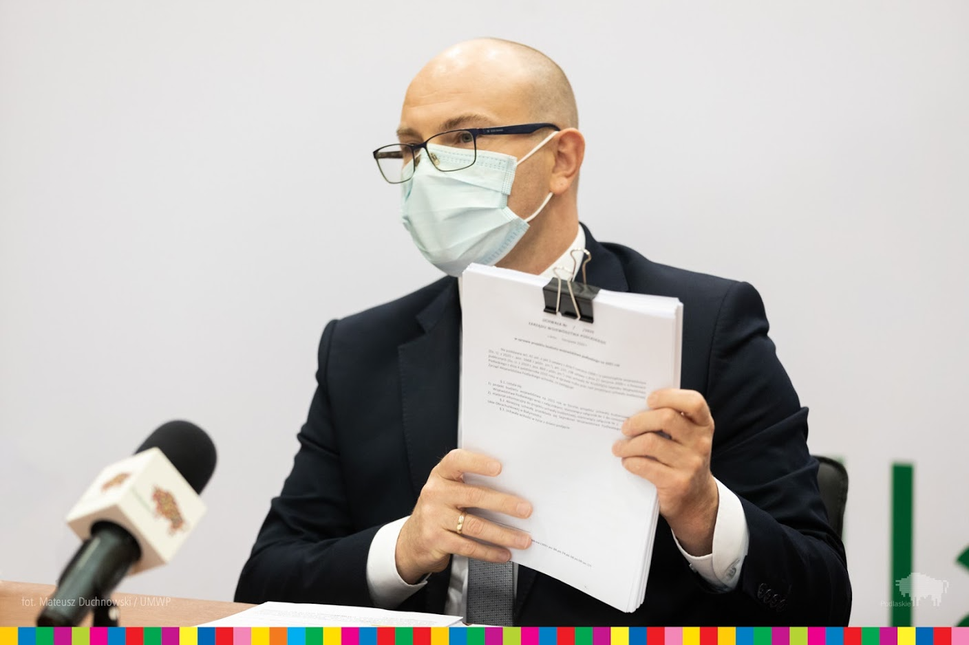 Marszałek siedzi na tle białej ściany, w rękach trzyma dokumenty spięte dużym czarnym spinaczem. Przed marszałkiem stoi mikrofon