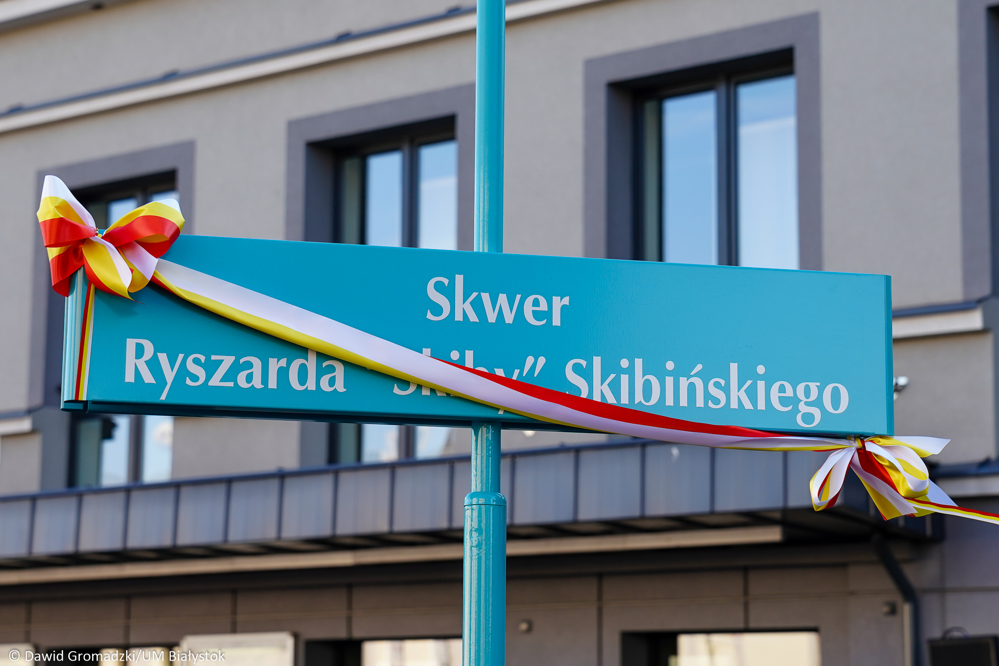 Niebieska tabliczka z nazwą skweru ryszarda skiby skibińskiego przepasana trójkolorową kokardą