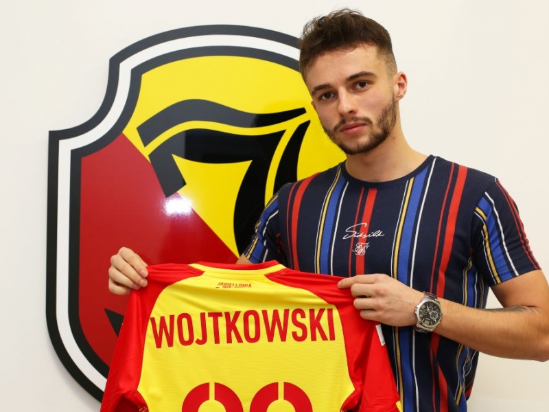 Mężczyzna trzyma klubową koszulkę Jagiellonii. Za jego plecami na ścianie wisi logo klubu.