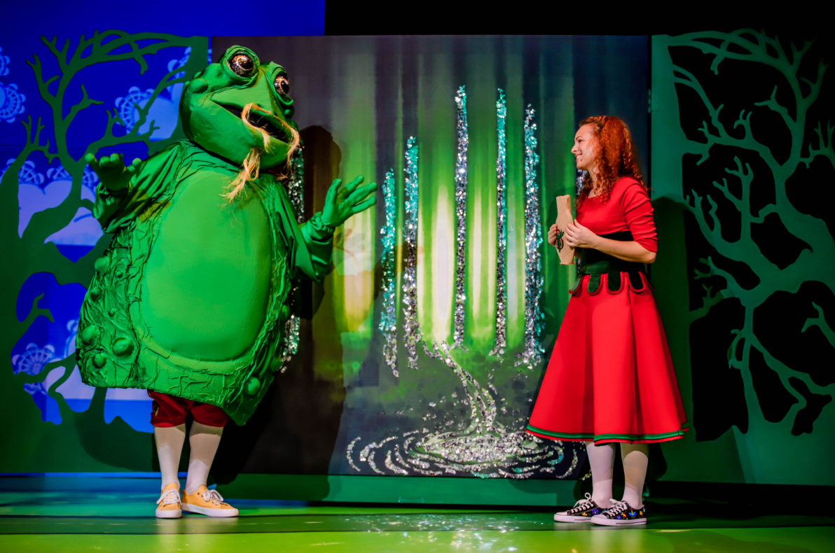 Na scenie teatralnej stoi aktor przebrany w dużą, zieloną żabę, obok stoi rudowłosa aktorka ubrana w czerwoną sukienkę