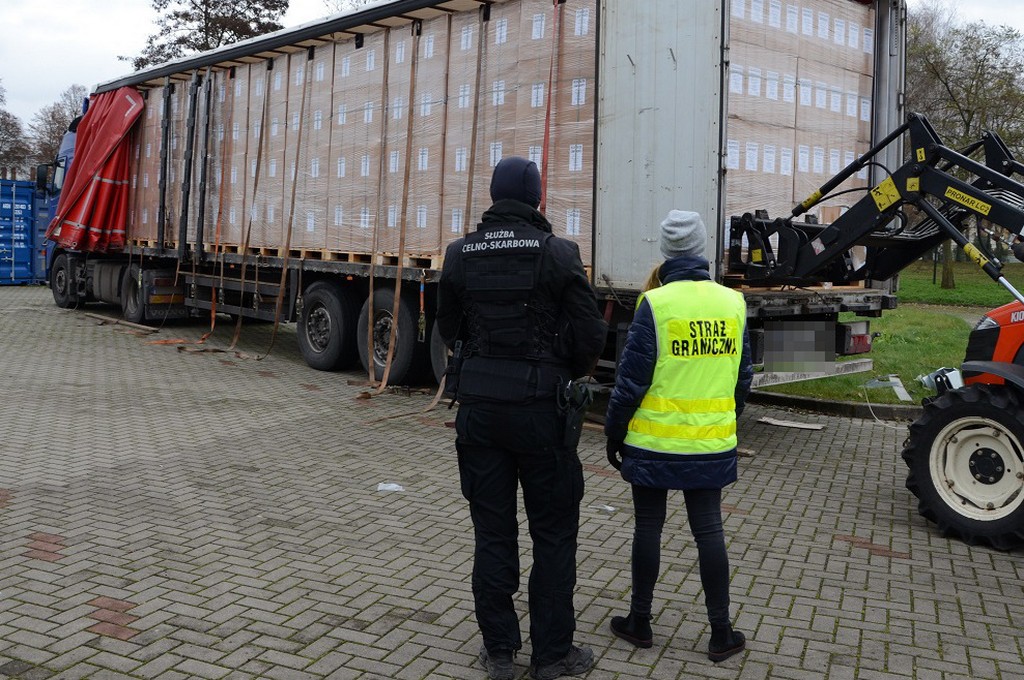 Ciężarówka wypchana nielegalnymi papierosami. Tirowi przygląda się dwóch funkcjonariuszy, stojący po lewej stronie jest ubrany na czarno, a osoba po prawej stronie ma na sobie zieloną odblaskową kamizelkę