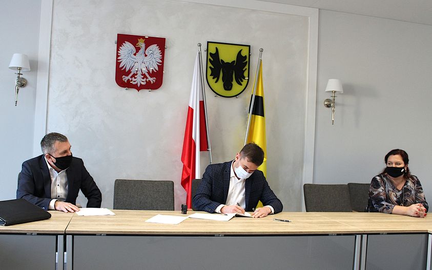 Przy stole siedzą trzy osoby, po środku burmistrz Wasilkowa, który podpisuje dokumenty. Za plecami osób siedzących przy stole jest godło Polski i Wasilkowa oraz flagi: Polski i Wasilkowa