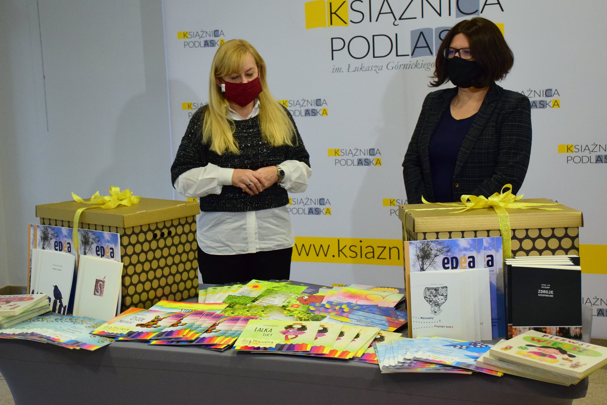 Dwie kobiety stoją za stołem, na którym ułożone są książki. Kobieta po lewej stronie ma długie, blond włosy i jest ubrane w czarne spodnie, białą koszulę i czarny sweterek. Kobieta po prawej stronie ma krótkie ciemne włosy, jest również ubrana na czarno. Za kobietami stoi baner z napisem Książnica Podlaska