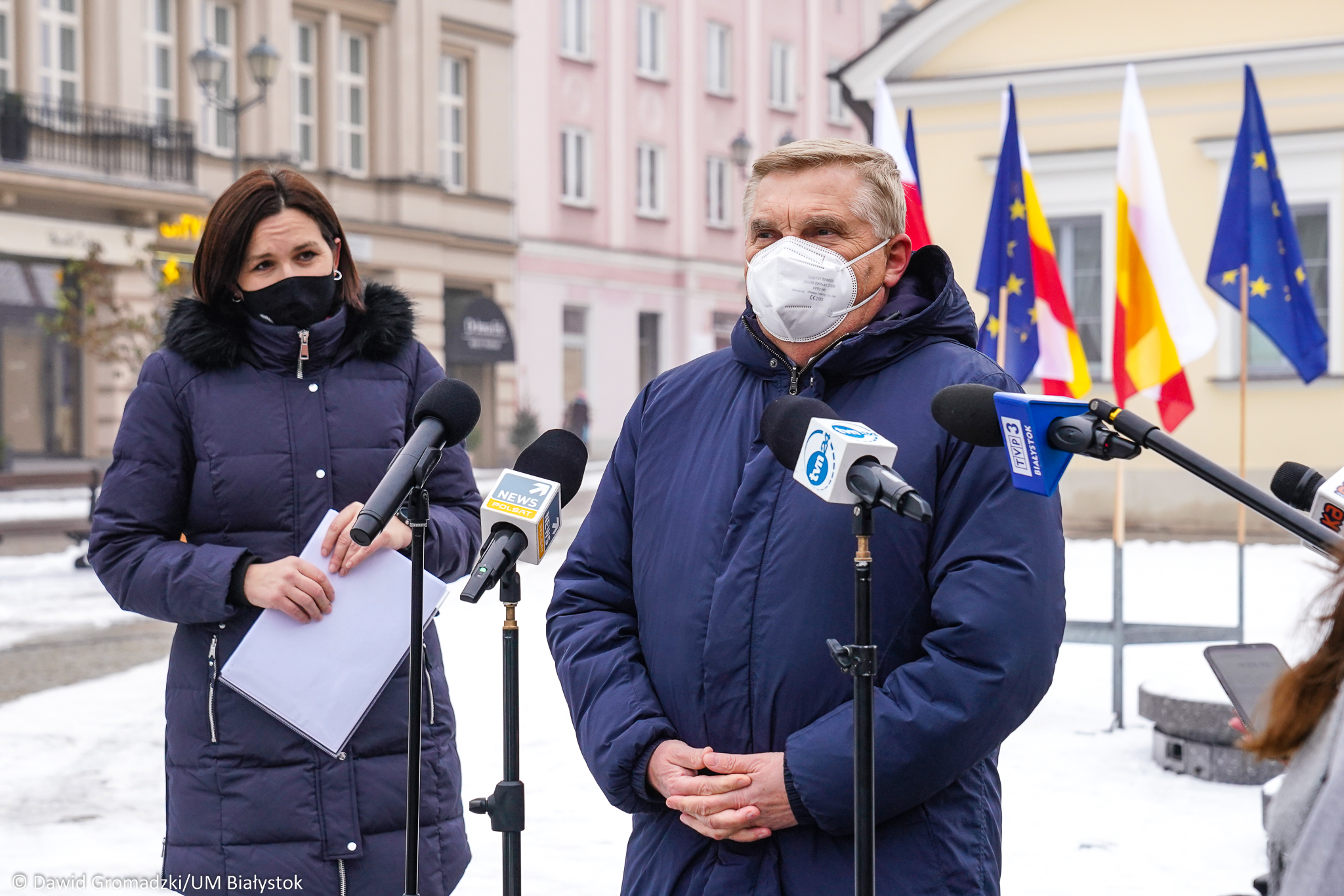 Prezydent Białegostoku stoi na Rynku Kościuszki podczas konferencji prasowej. Mówi do mikrofonów dziennikarskich. Za prezydentem, po prawej stronie widoczne są flagi Unii Europejskiej, Białegostoku i Polski. Po lewej stronie stoi rzecznik prasowa prezydenta