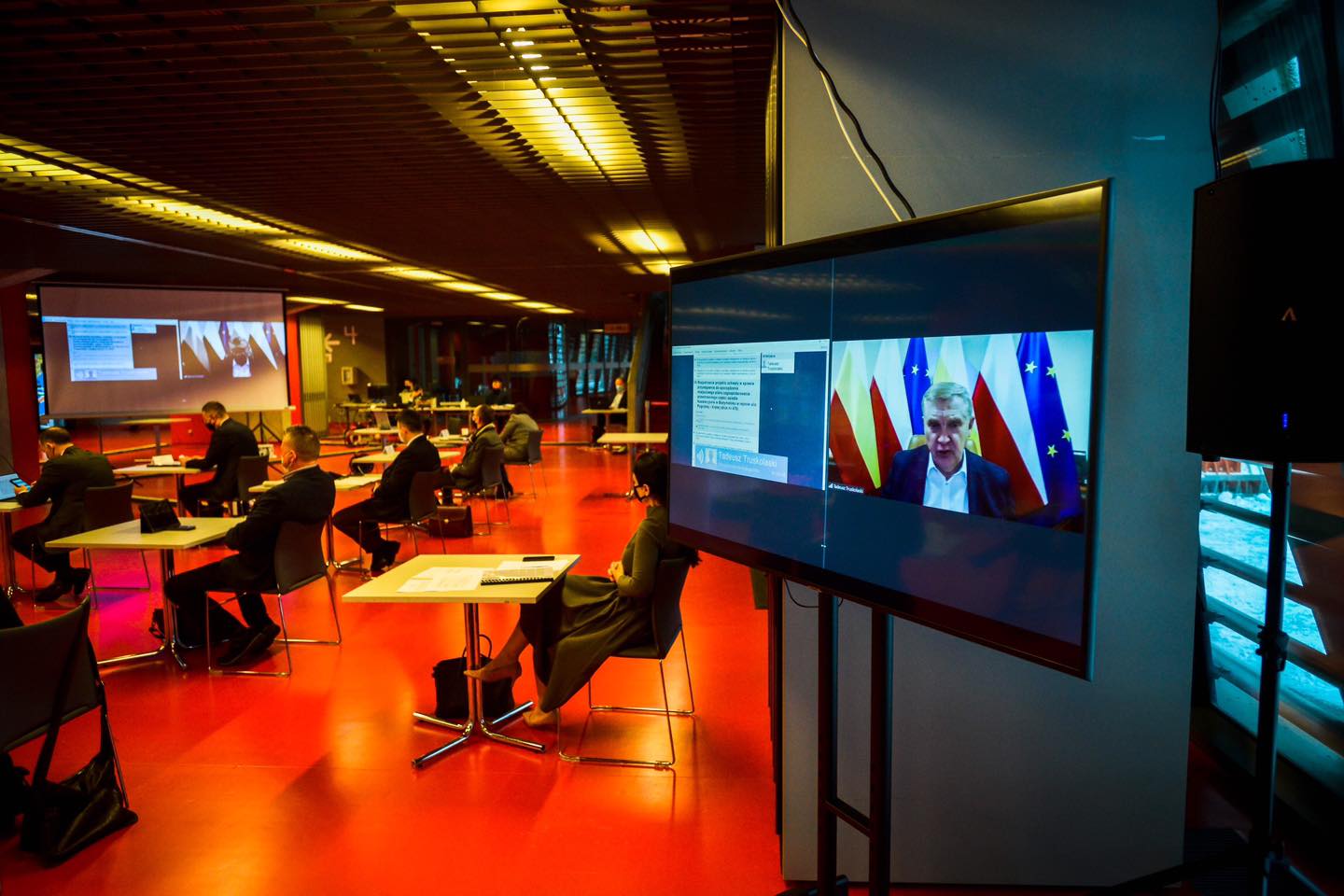 Sala konferencyjna Stadionu Miejskiego w Białymstoku. Po lewej stronie, przy pojedynczych stolikach siedzą radni, po prawej stronie jest ustawiony duży monitor, na którym widoczny jest prezydent Białegostoku