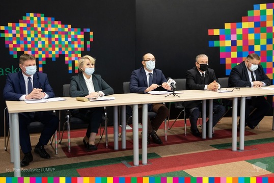 Przy stole, obok siebie, siedzi pięć osób, jedna kobieta, czterech mężczyzn. Za ich plecami jest czarna ściana, na której jest logo województwa podlaskiego: różnokolorowy, pikselowy żubr