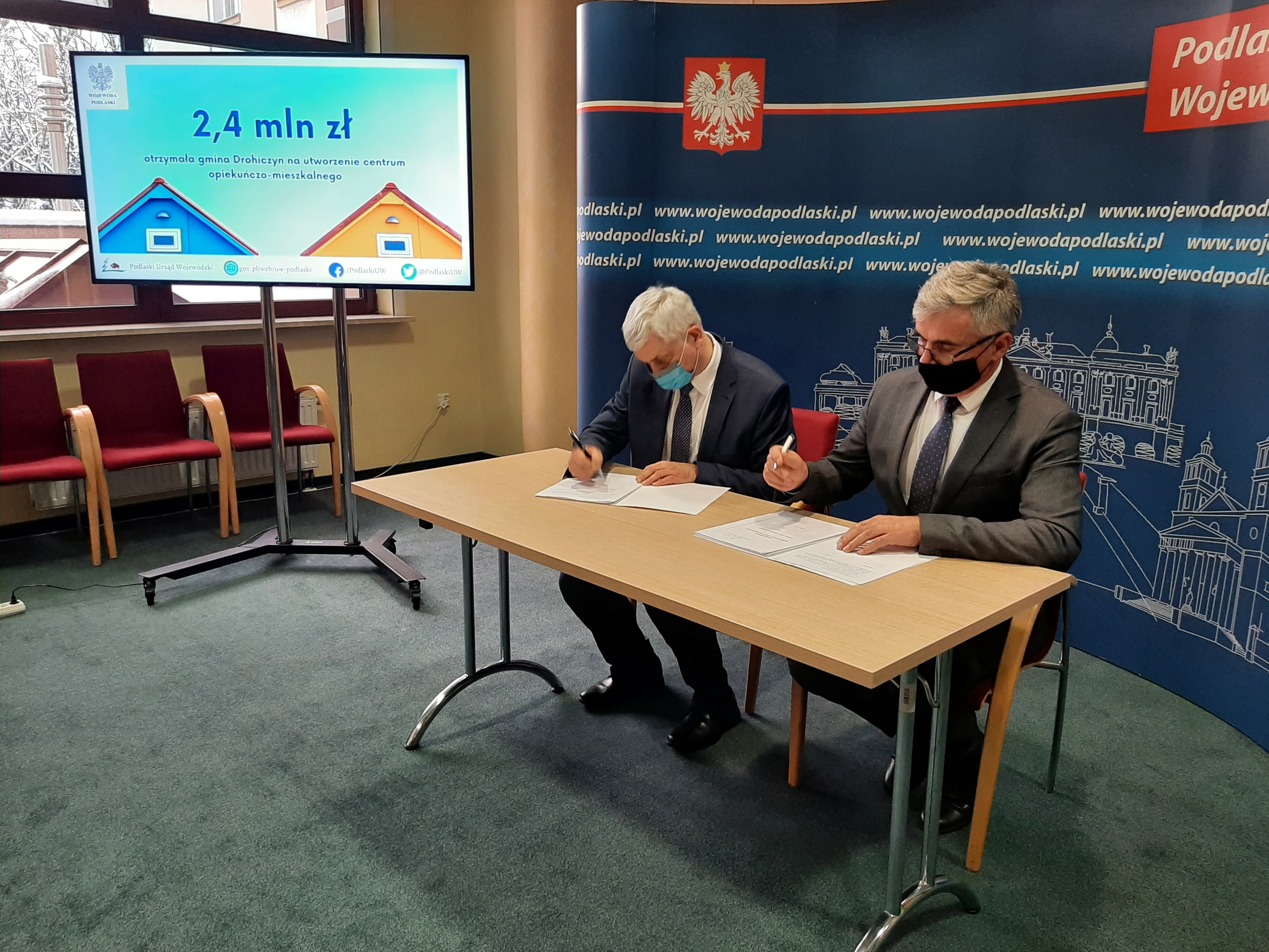 Wojewoda i burmistrz gminy Drohiczyn podpisują umowę. Mężczyźni siedzą przy stole, za nimi znajduje się ścianka Podlaskiego Urzędu Wojewódzkiego w Białymstoku. W lewym rogu zdjęcia widoczny jest duży telewizor, na którym jest wyświetlona kwota 2.4 miliona złotych.