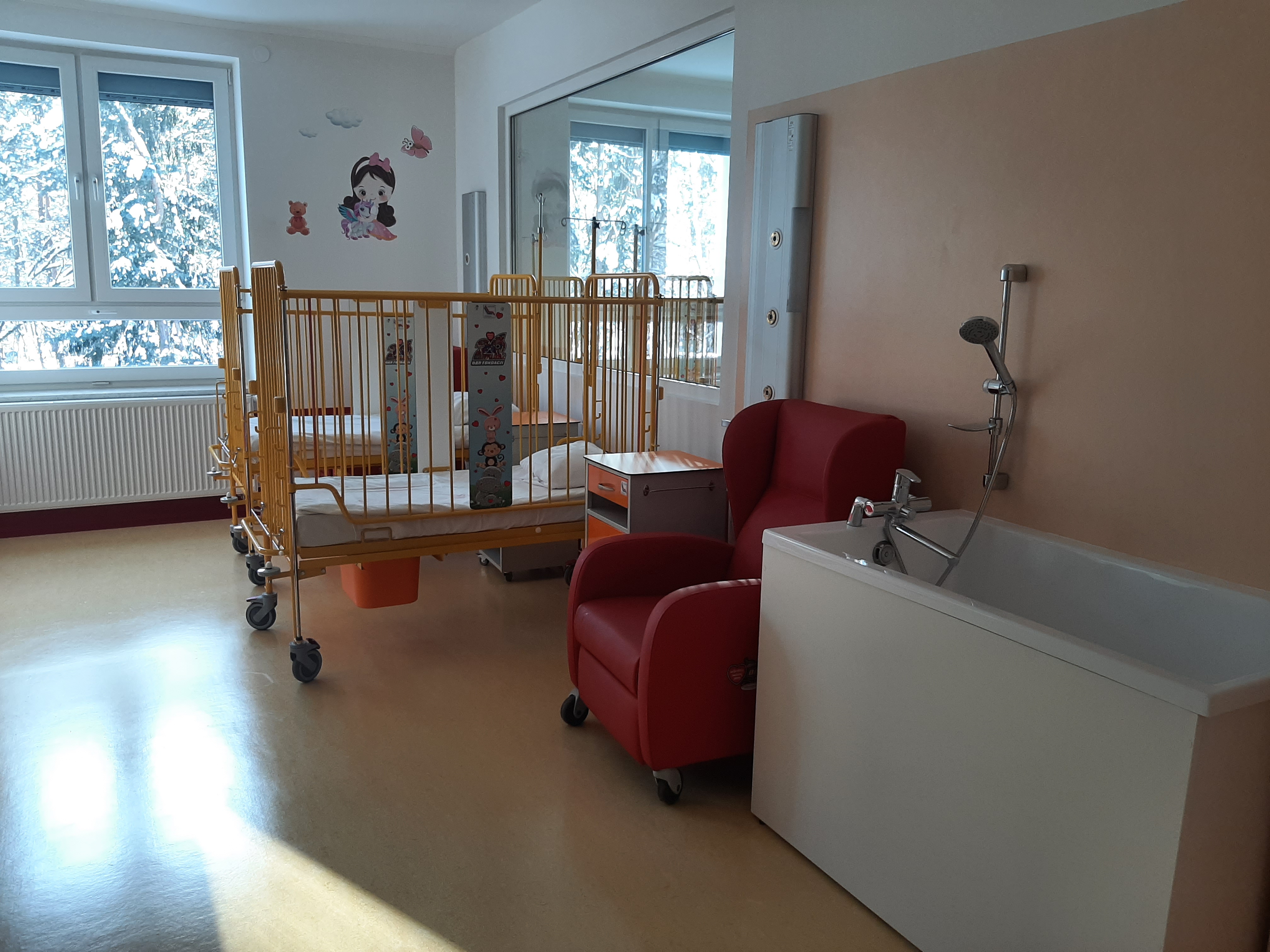 Sala szpitalna, pod ścianą, po prawej stronie stoi wanna, czerwony fotel i żółte łóżeczka ze szczebelkami dla małych dzieci. Po lewej stronie znajduje się okno