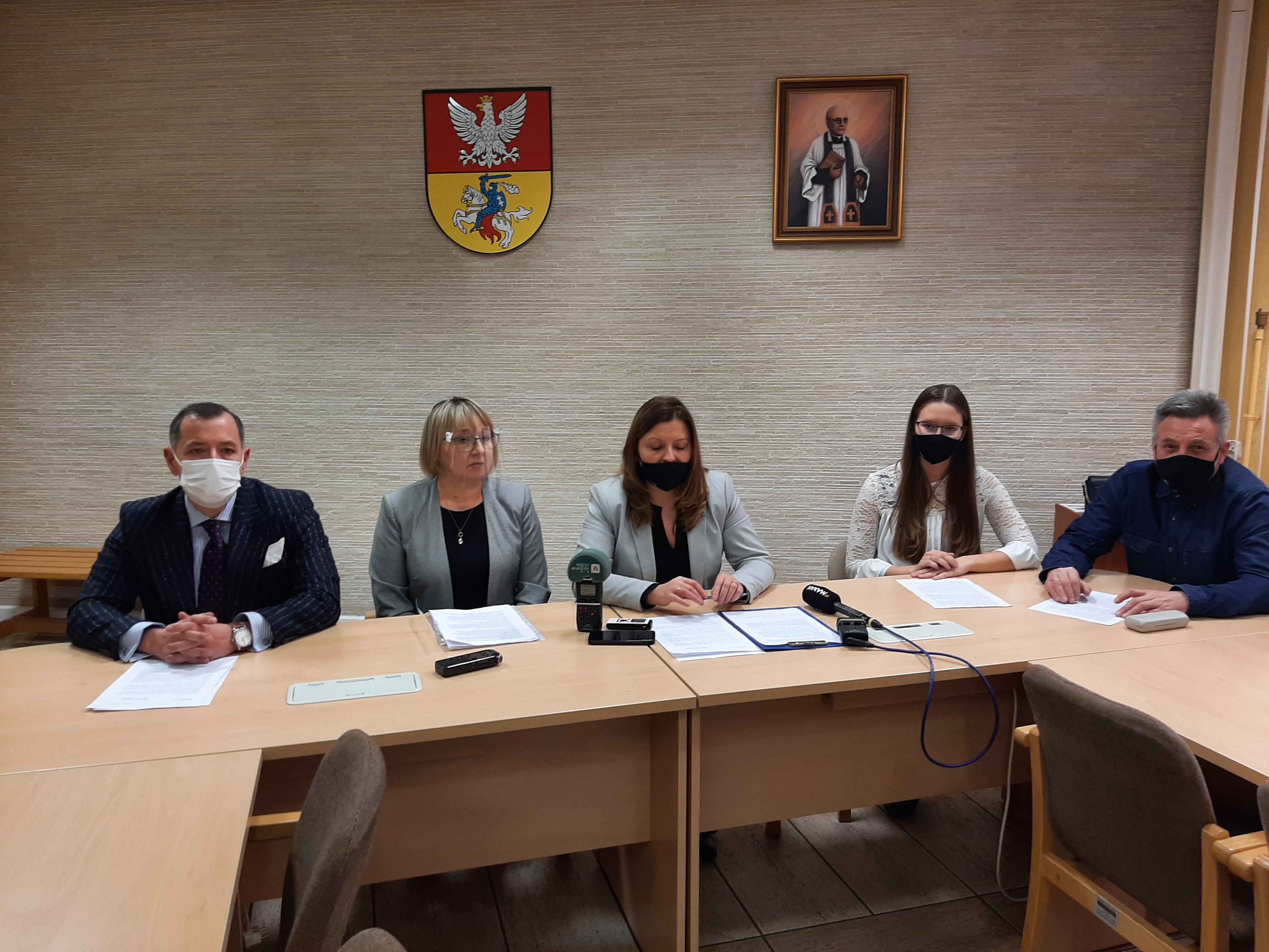 Pięciu radnych siedzi przy stole, po środku kobiety, bo bokach mężczyźni. Za nimi na ścianie wisi herb Białegostoku oraz wizerunek patrona Białegostoku błogosławionego księdza Michała Sopoćki