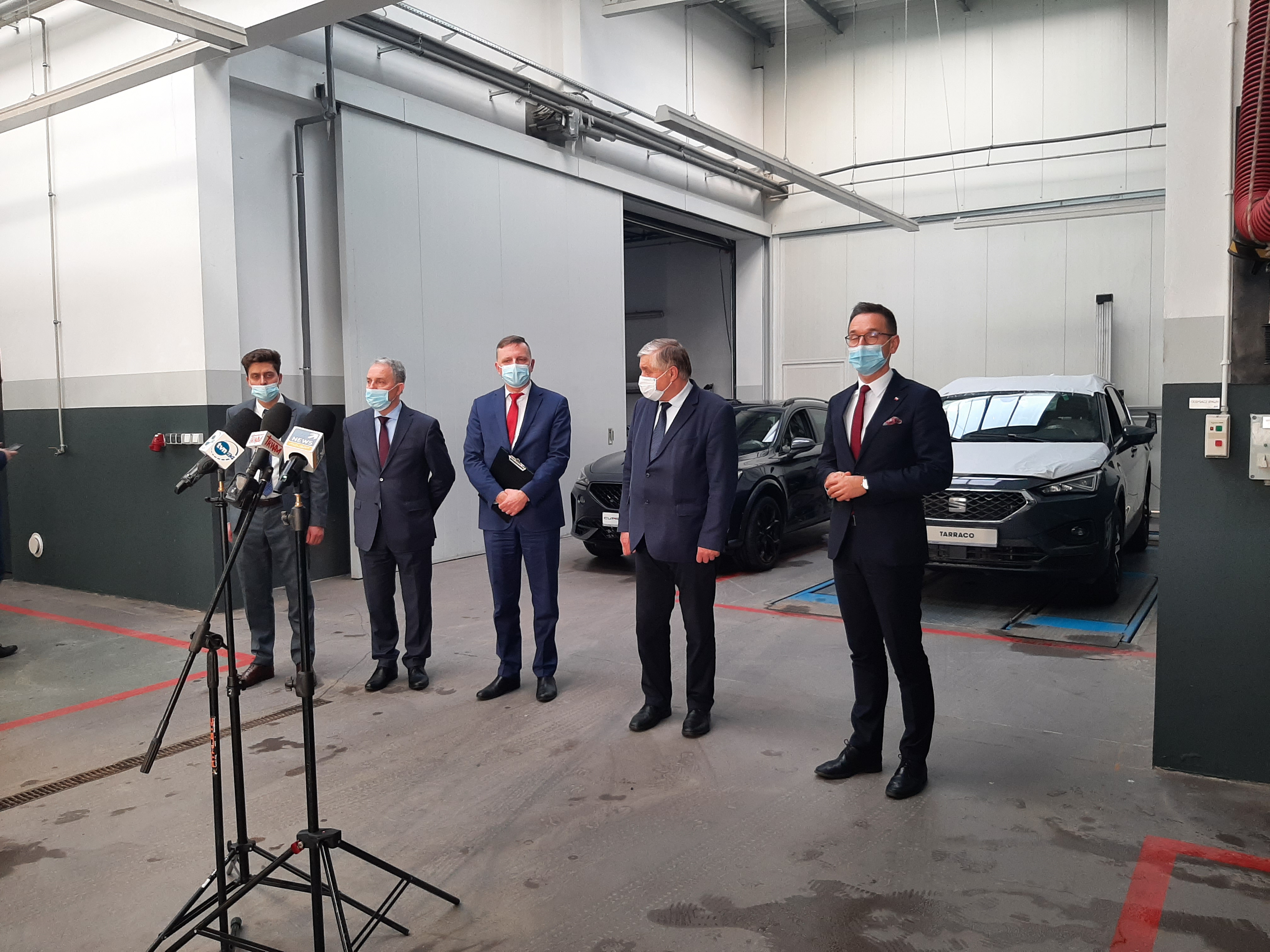 Pięciu mężczyzn stoi w hali, w której serwisowane i naprawiane są samochody. Za nimi stoją dwa uta, natomiast przed nimi mikrofony, po trwa konferencja prasowa