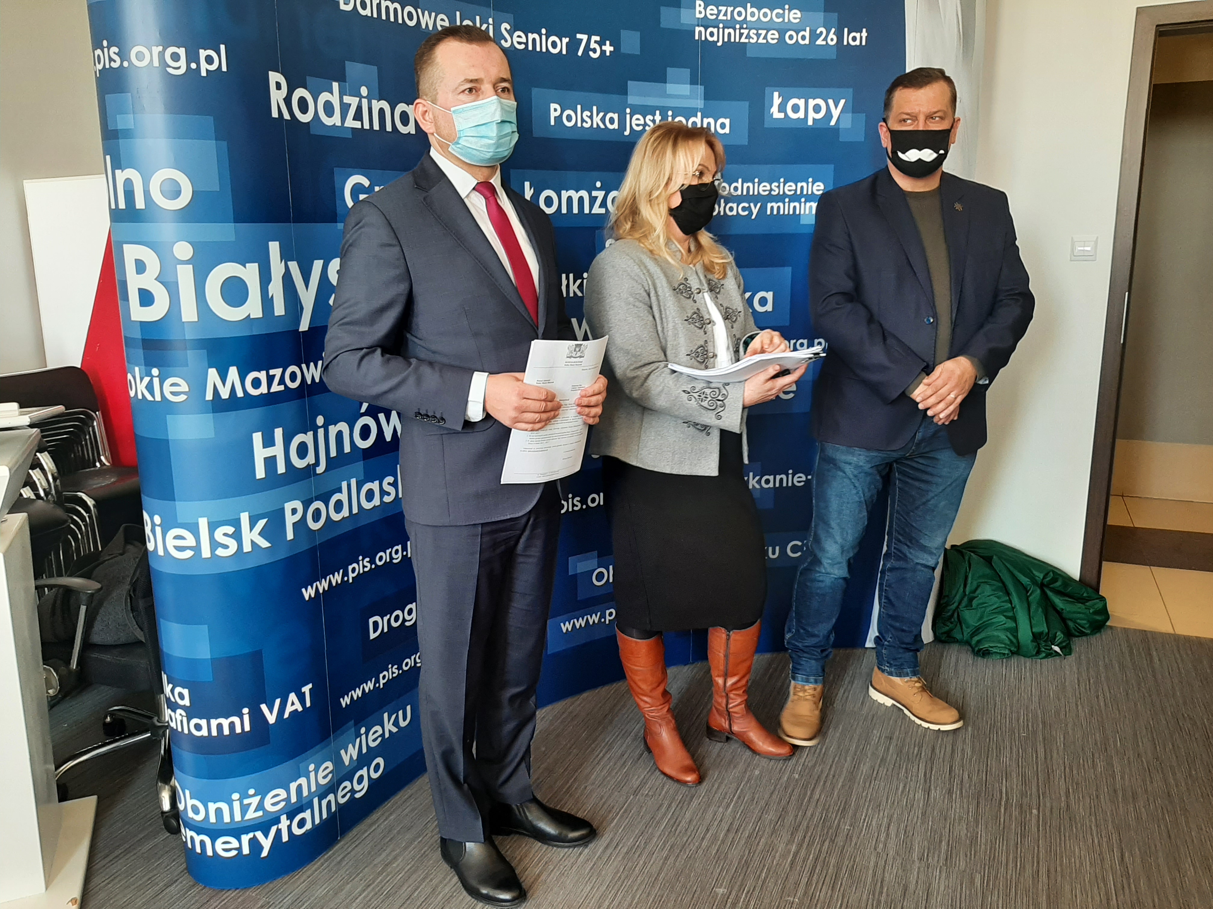 Na niebieskiej ściance w różne białe napisy stoją trzy osoby, po środku jest kobieta