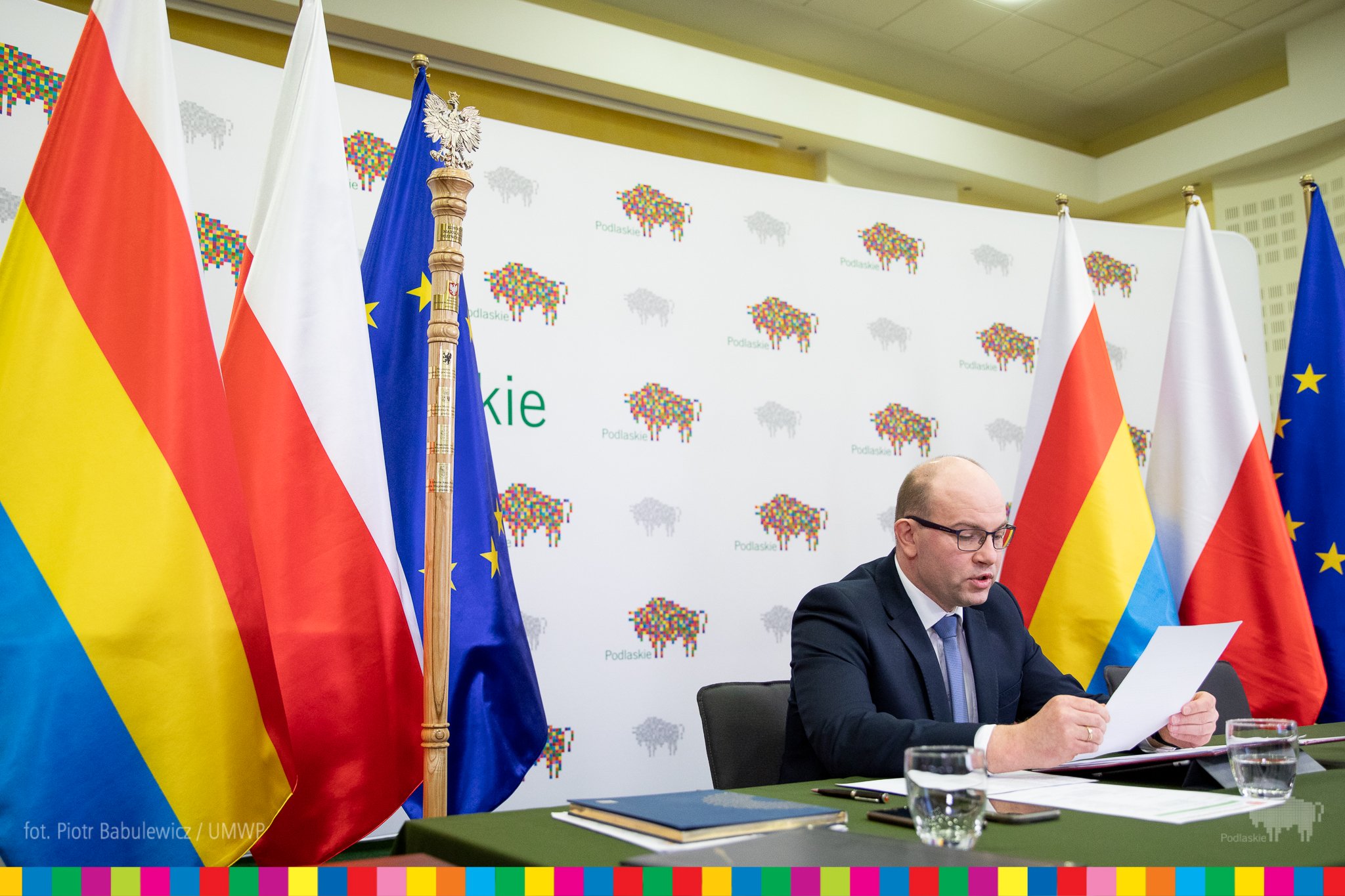 Marszałek siedzi przy stole nakrytym zielonym materiałem, za nim znajduje się ścianka, w logo - żubra, województwa podlaskiego. Po obu stronach marszałka znajdują się flagi województwa podlaskiego, Polski, Unii Europejskiej